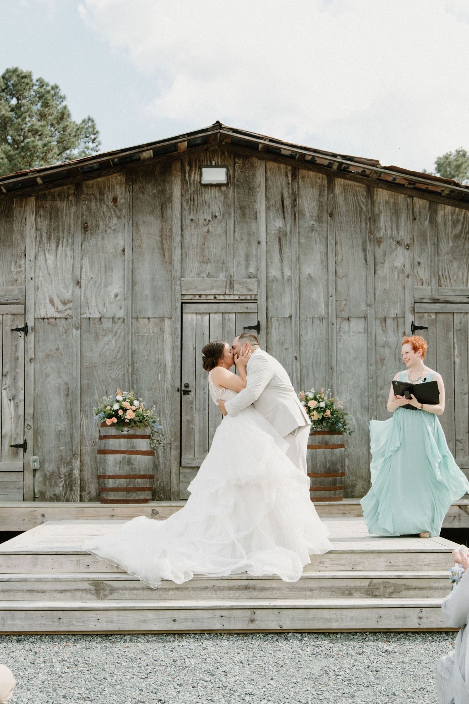 A rustic wedding ceremony takes place on a wooden deck in front of a weathered barn with floral barrel arrangements.