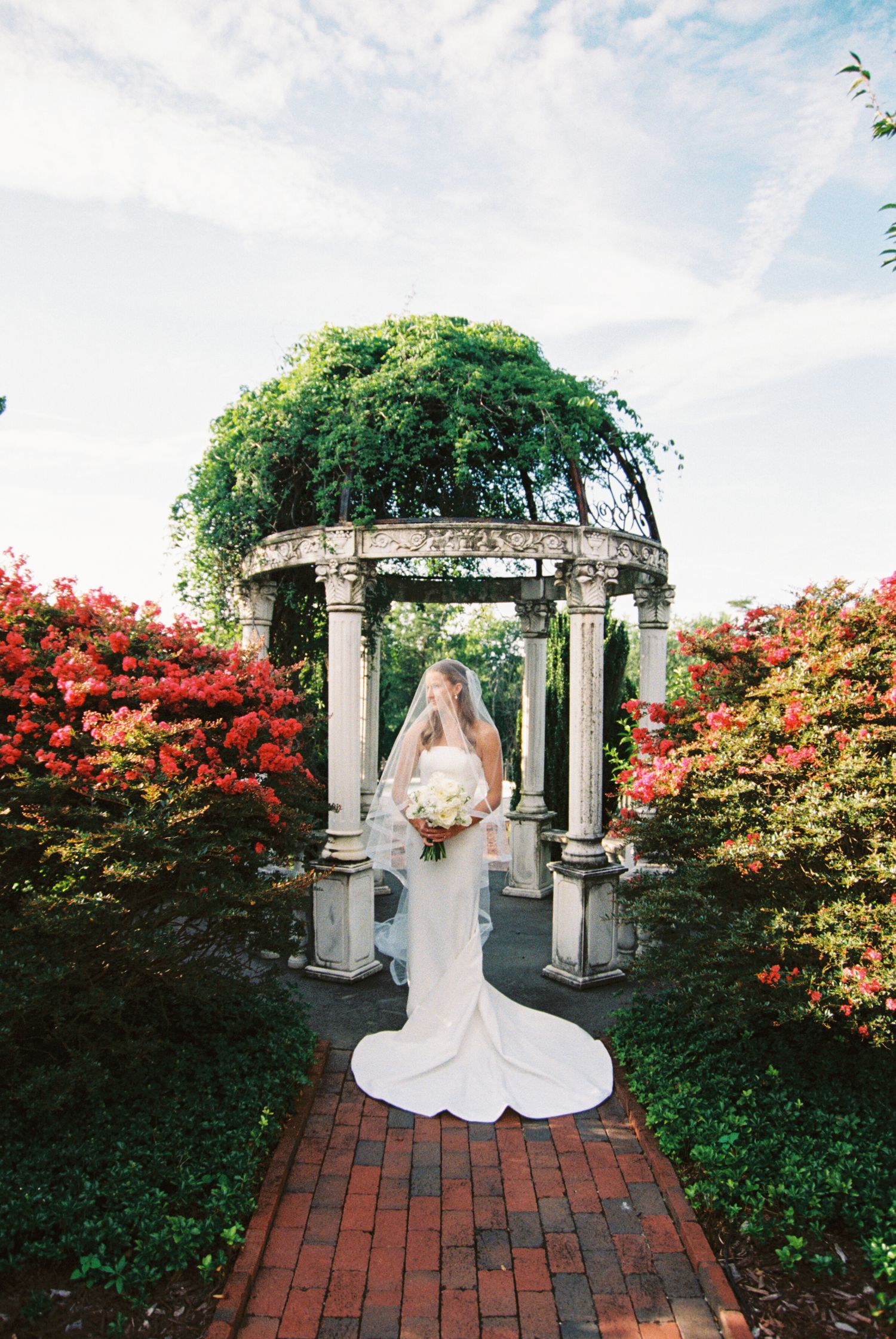 Bride in white gown and veil stands before a vine-covered gazebo surrounded by red azaleas on a brick path.