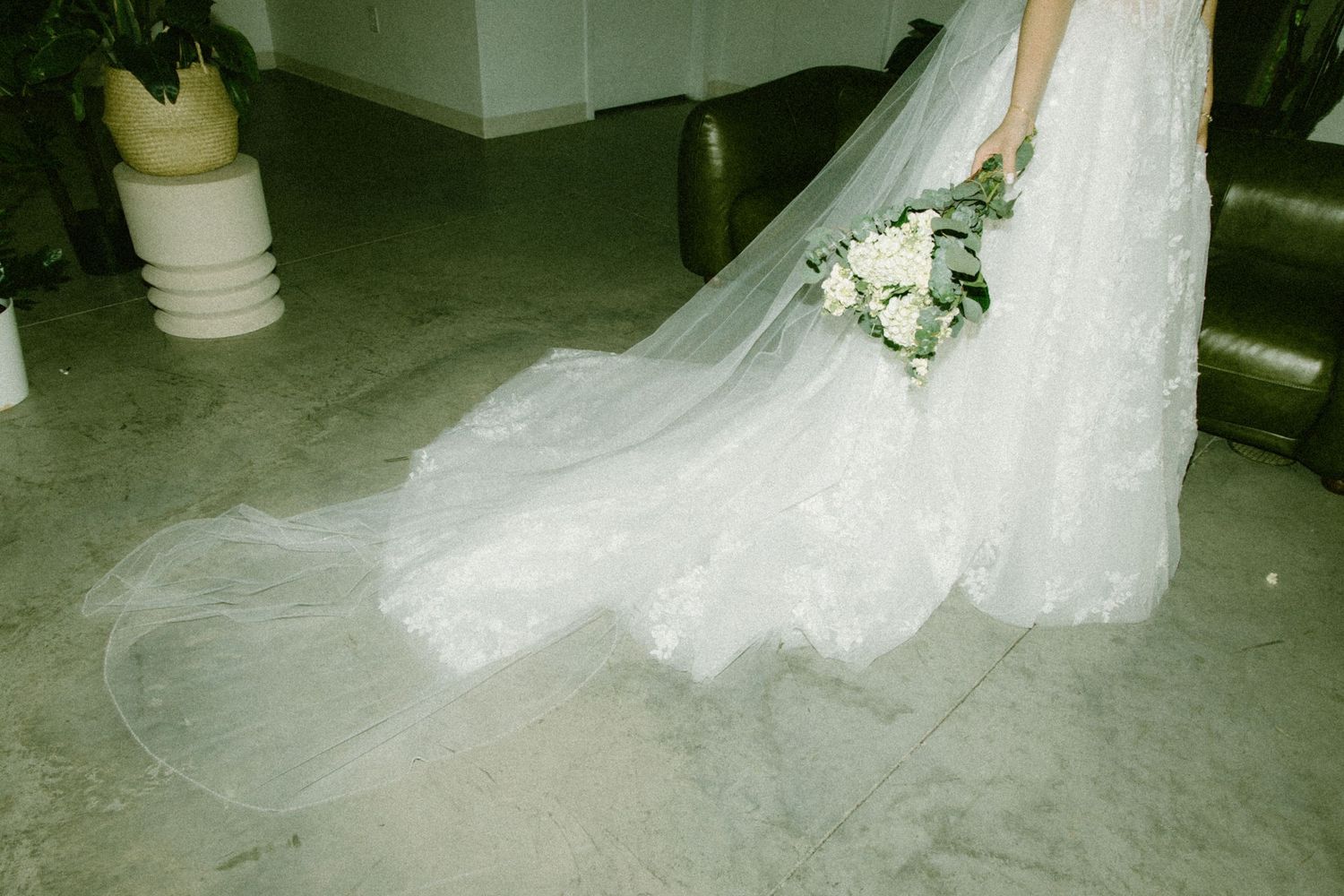 A flowing white wedding dress with delicate lace train and white floral bouquet rests on a concrete floor.