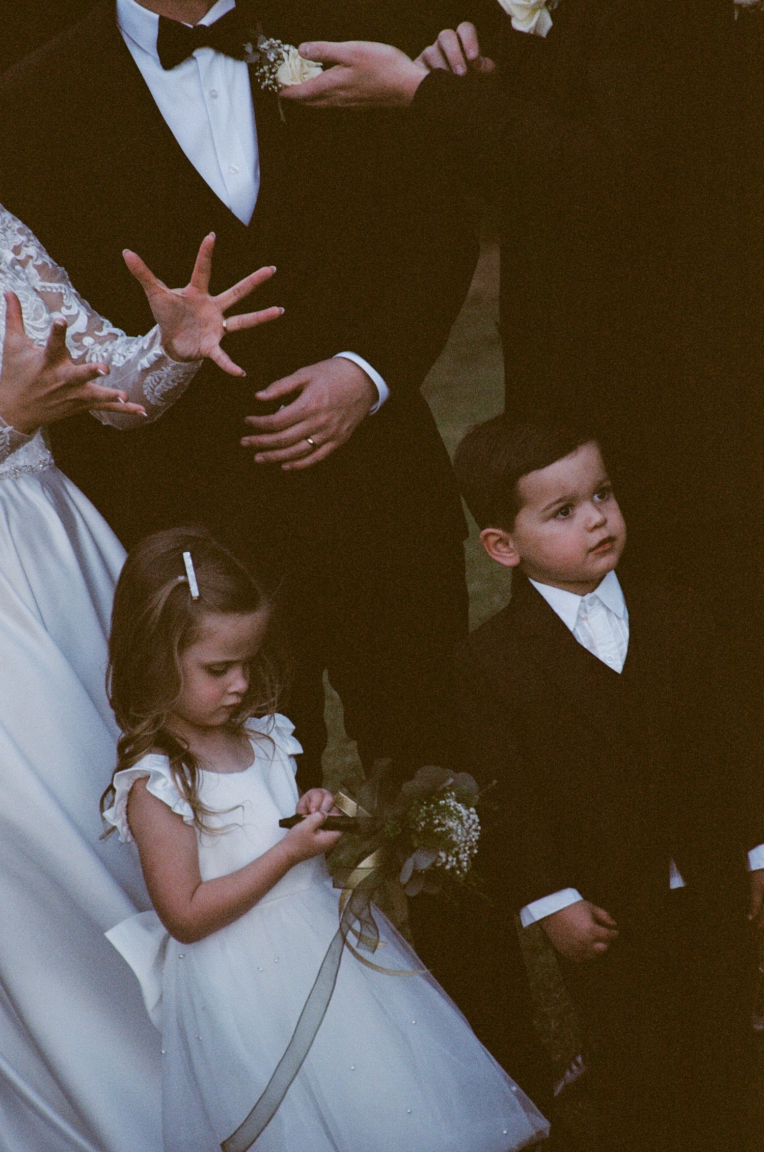 A small flower girl in a white dress stands among formally dressed guests at a wedding ceremony.