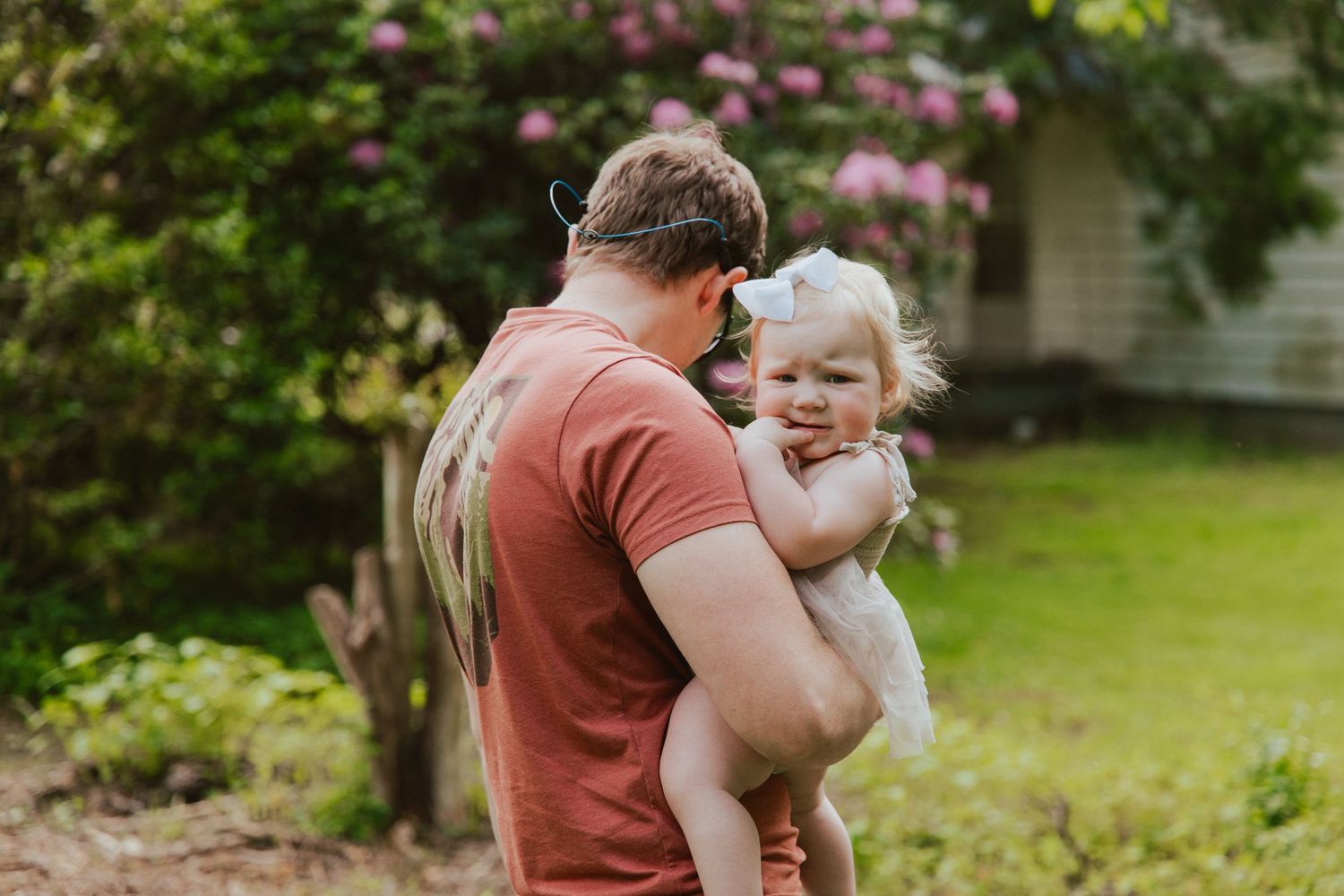 An adult in a rust-colored shirt holds a small child while walking in a lush garden on a sunny day.