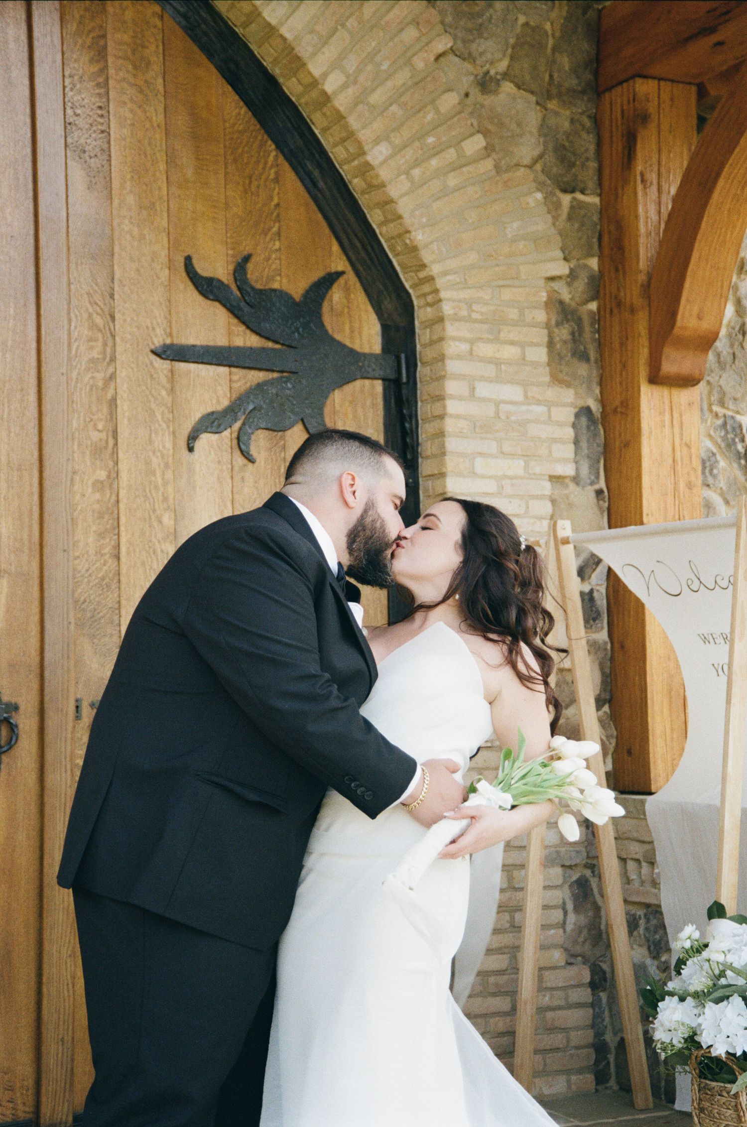 A romantic wedding moment between a couple kissing beneath a brick archway with a decorative metal sun detail.