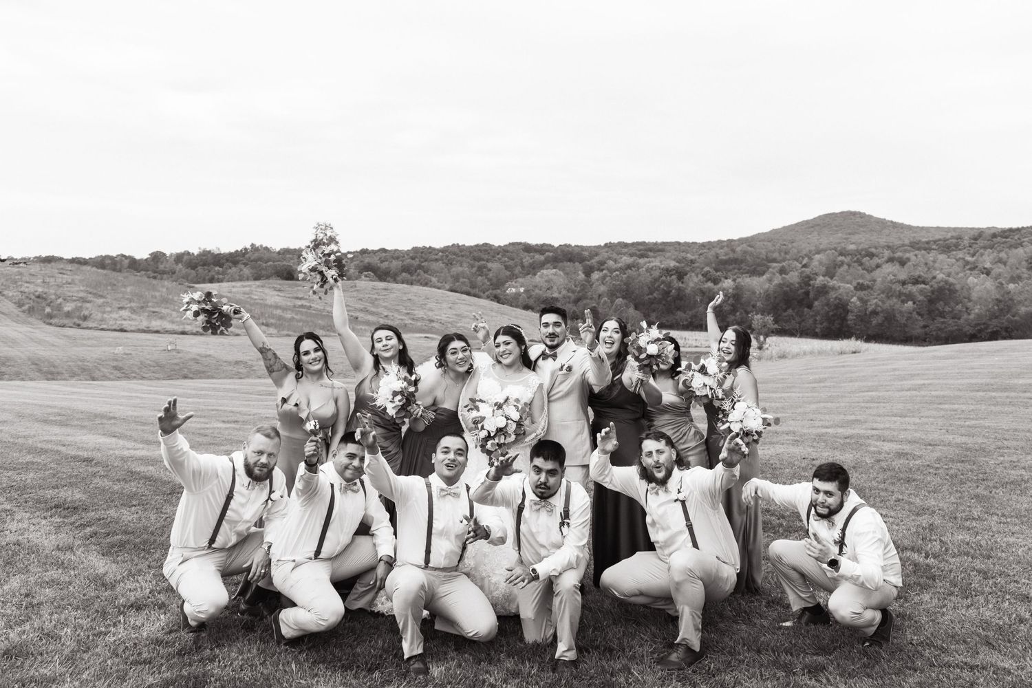 Wedding party in white and blue poses casually on a grassy hillside in black and white.
