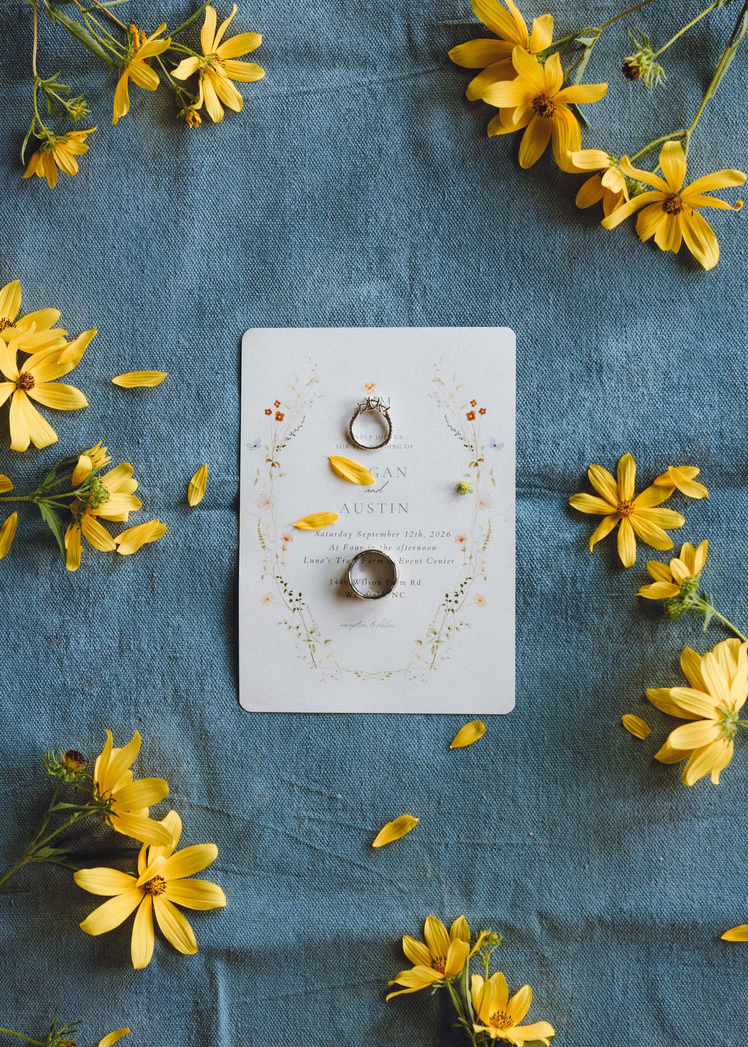 Wedding rings resting on floral invitation card surrounded by yellow daisy flowers on blue linen background.