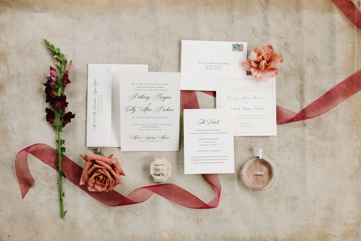 Wedding invitations and stationery arranged with dusty rose ribbons and floral accents on a neutral textured background.