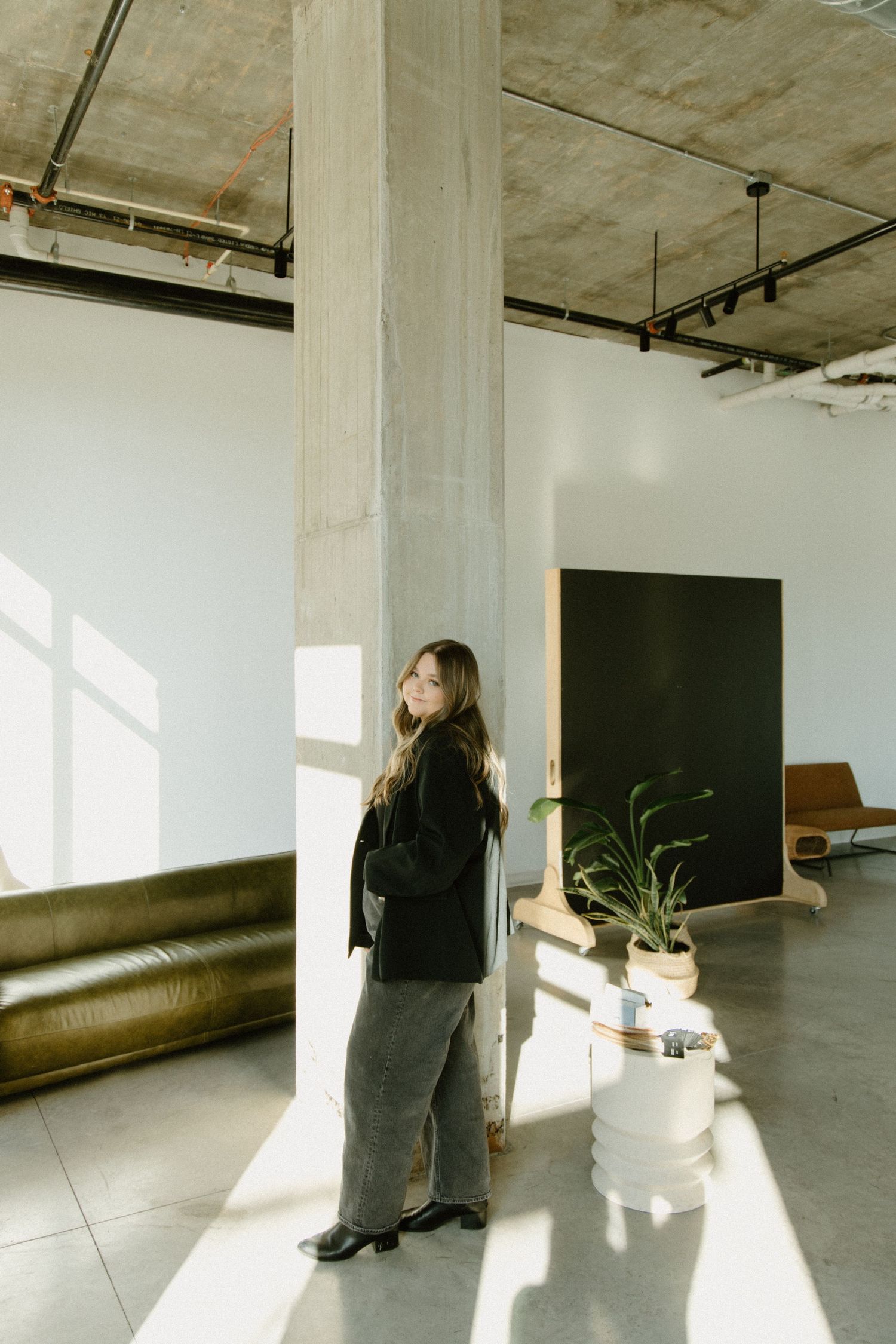 Minimalist industrial space with sunlit interior, concrete column, brown leather couch and potted plant.