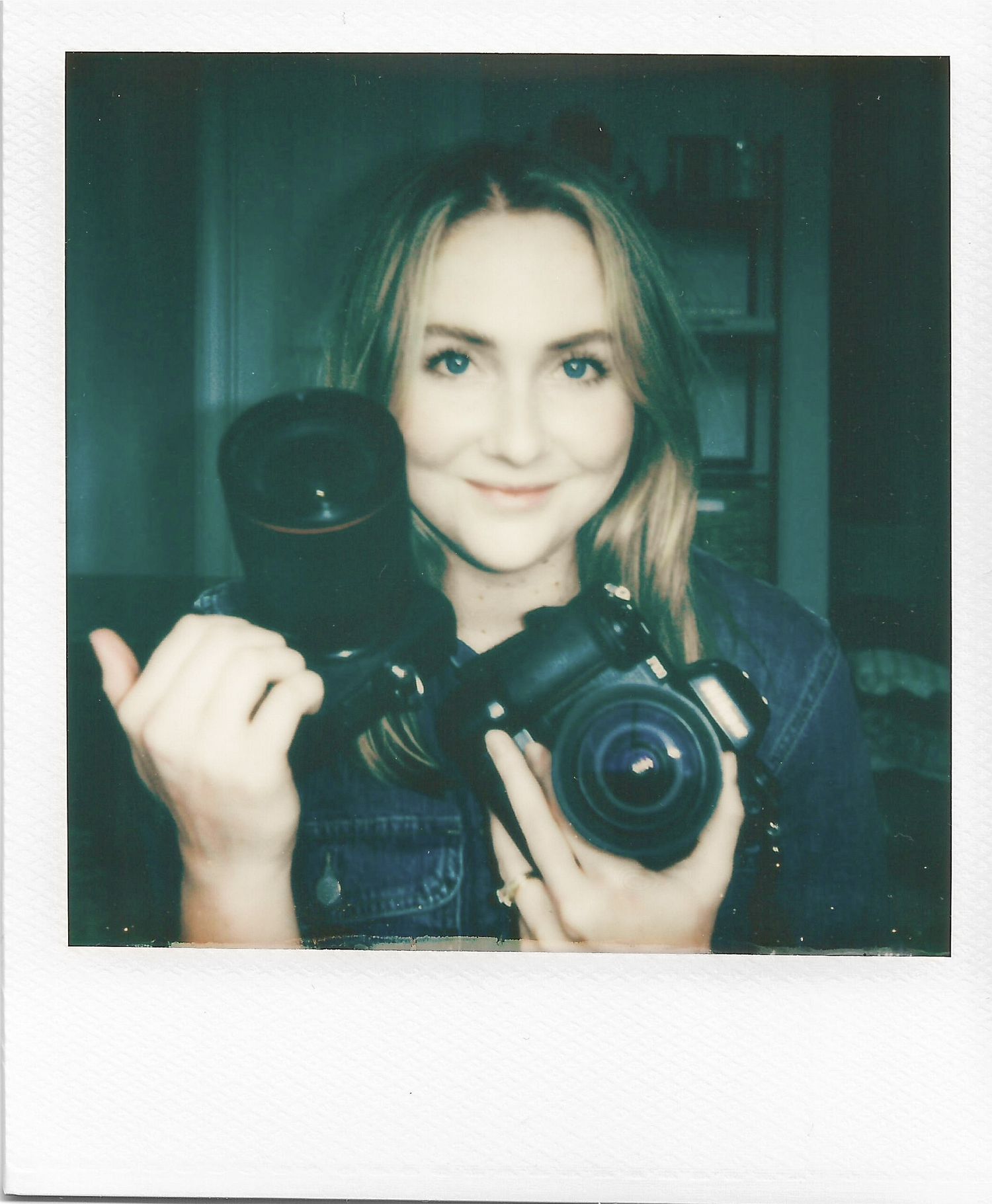 Polaroid photo shows photographer in dark top holding professional DSLR camera and lens against teal background.