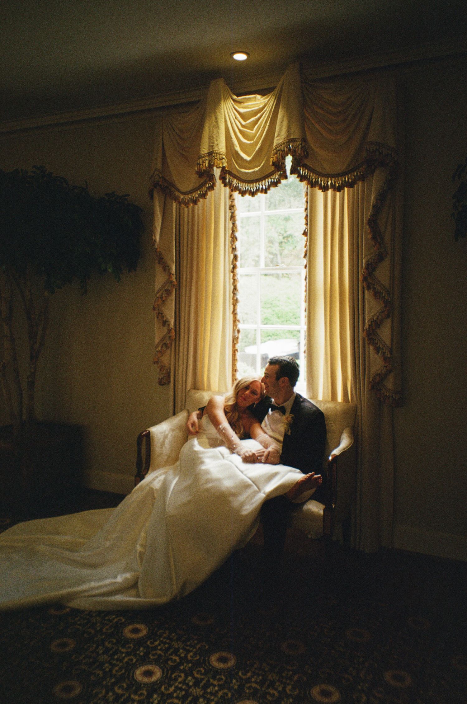 A romantic wedding couple shares intimate moments near a window in an elegant room with flowing curtains.