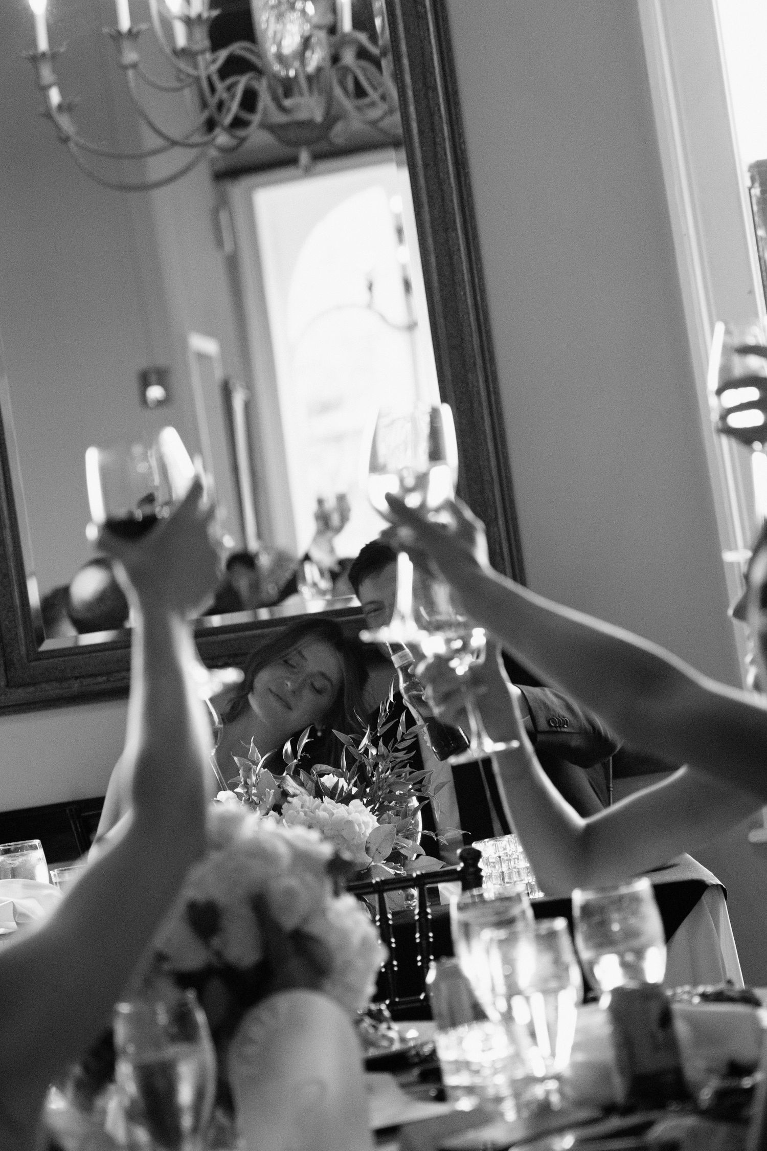 Black and white photo of people raising glasses in a toast at an elegant dining table with glassware and flowers.