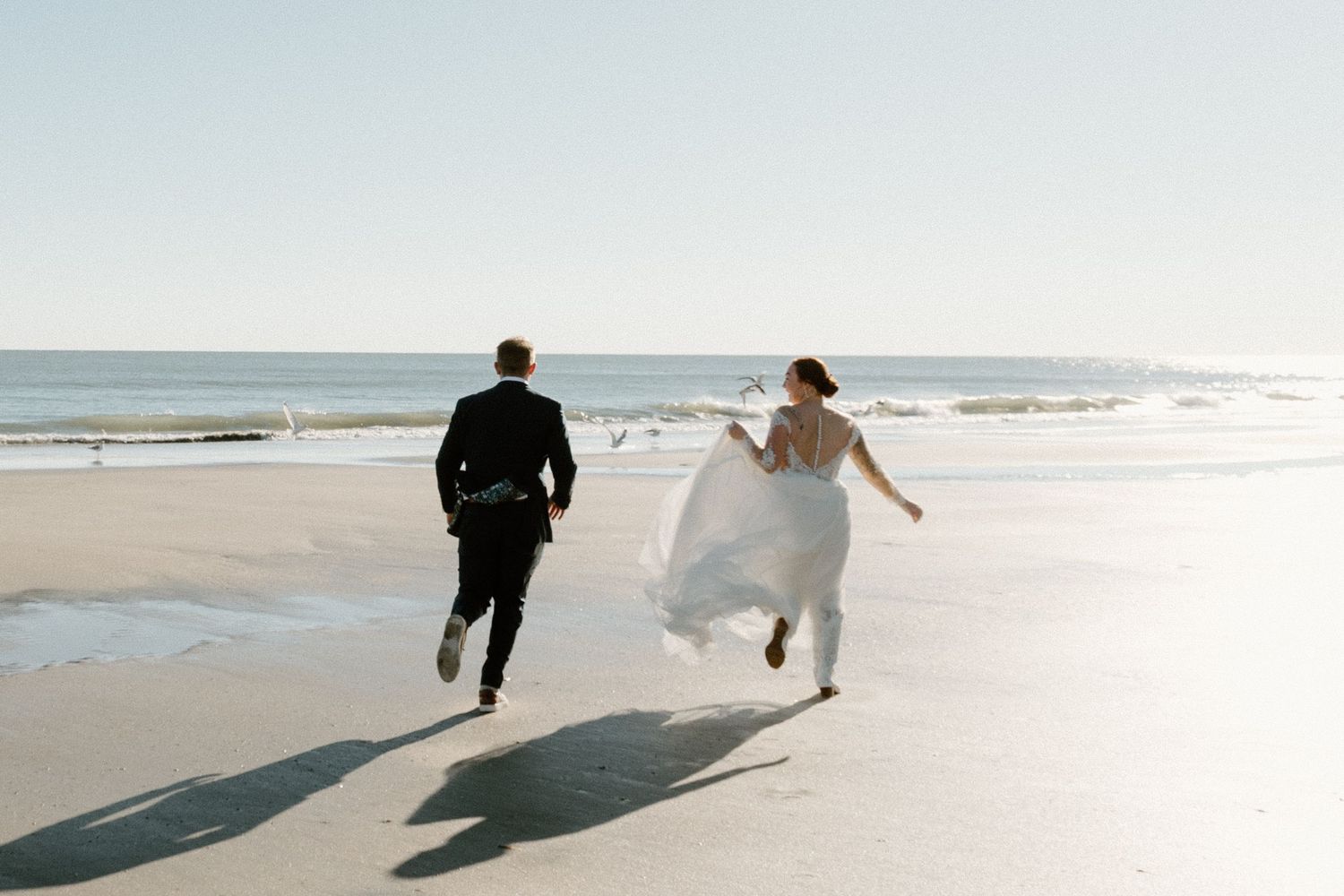 Two people in wedding attire run joyfully along a sunny beach with ocean waves in the background.