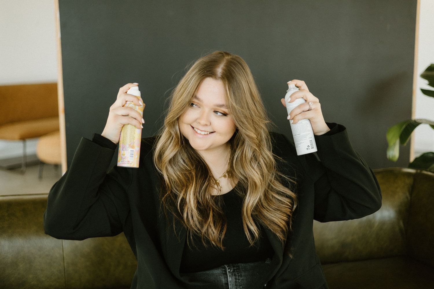 A woman in a black sweater smiles while holding up two hair spray bottles against a dark leather couch.