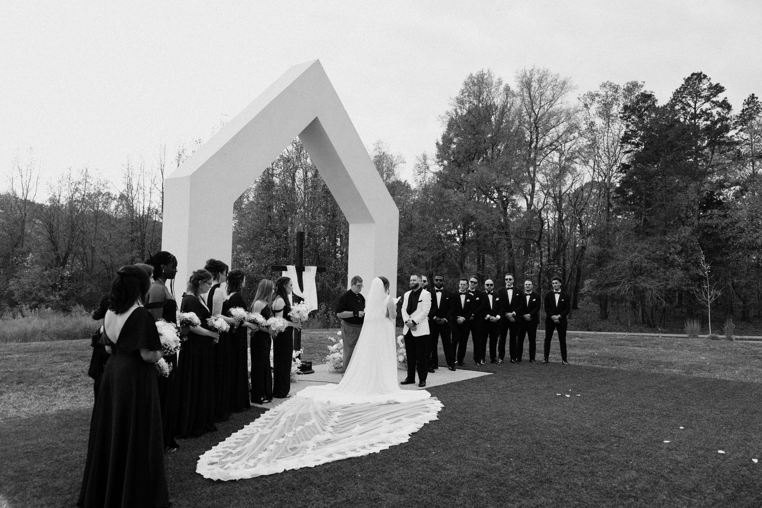 Black and white outdoor wedding ceremony with bride, groom, wedding party, and geometric arch on a grassy lawn.