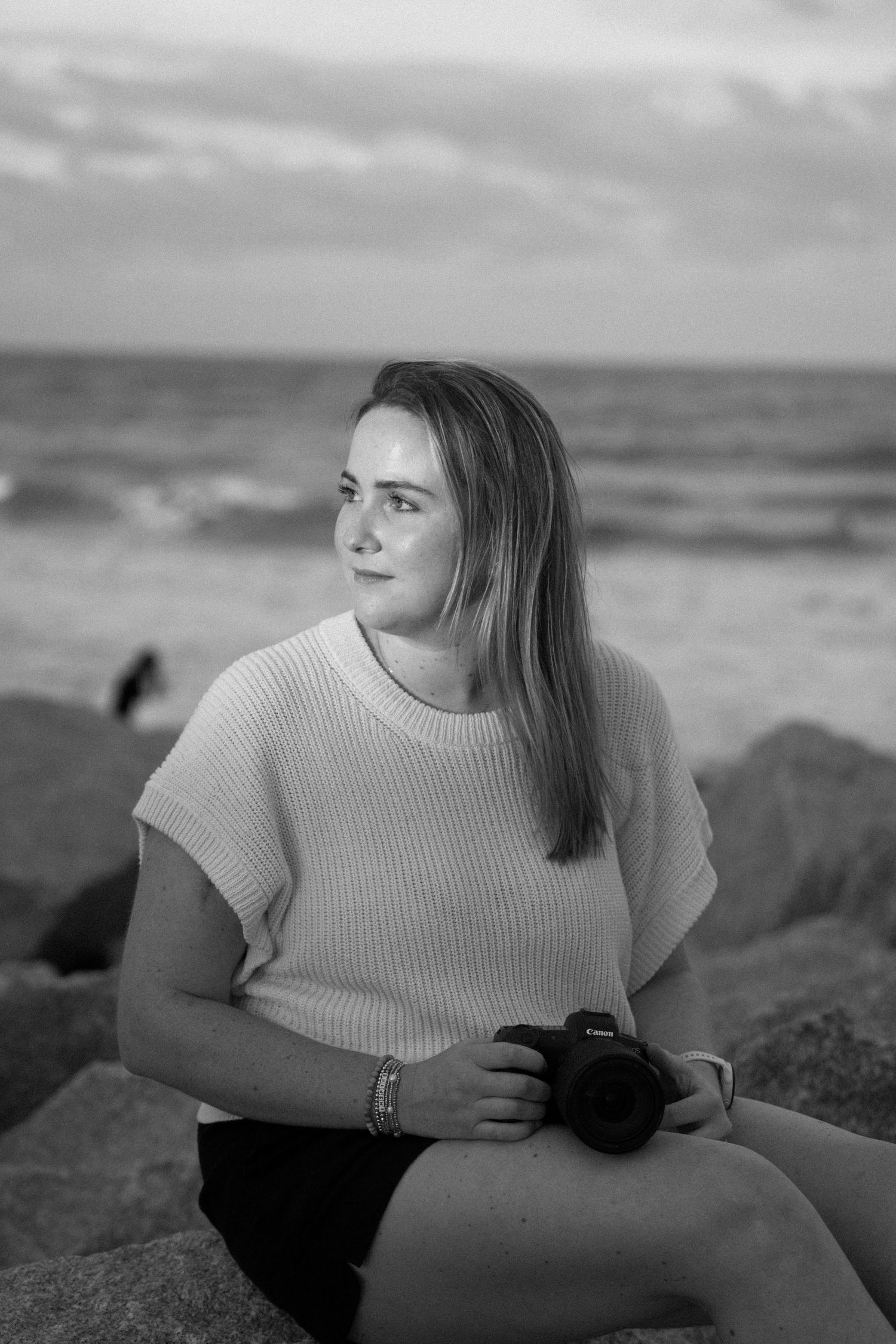 Black and white portrait series showing a person posing on a beach in a light colored sweater against a cloudy sky.