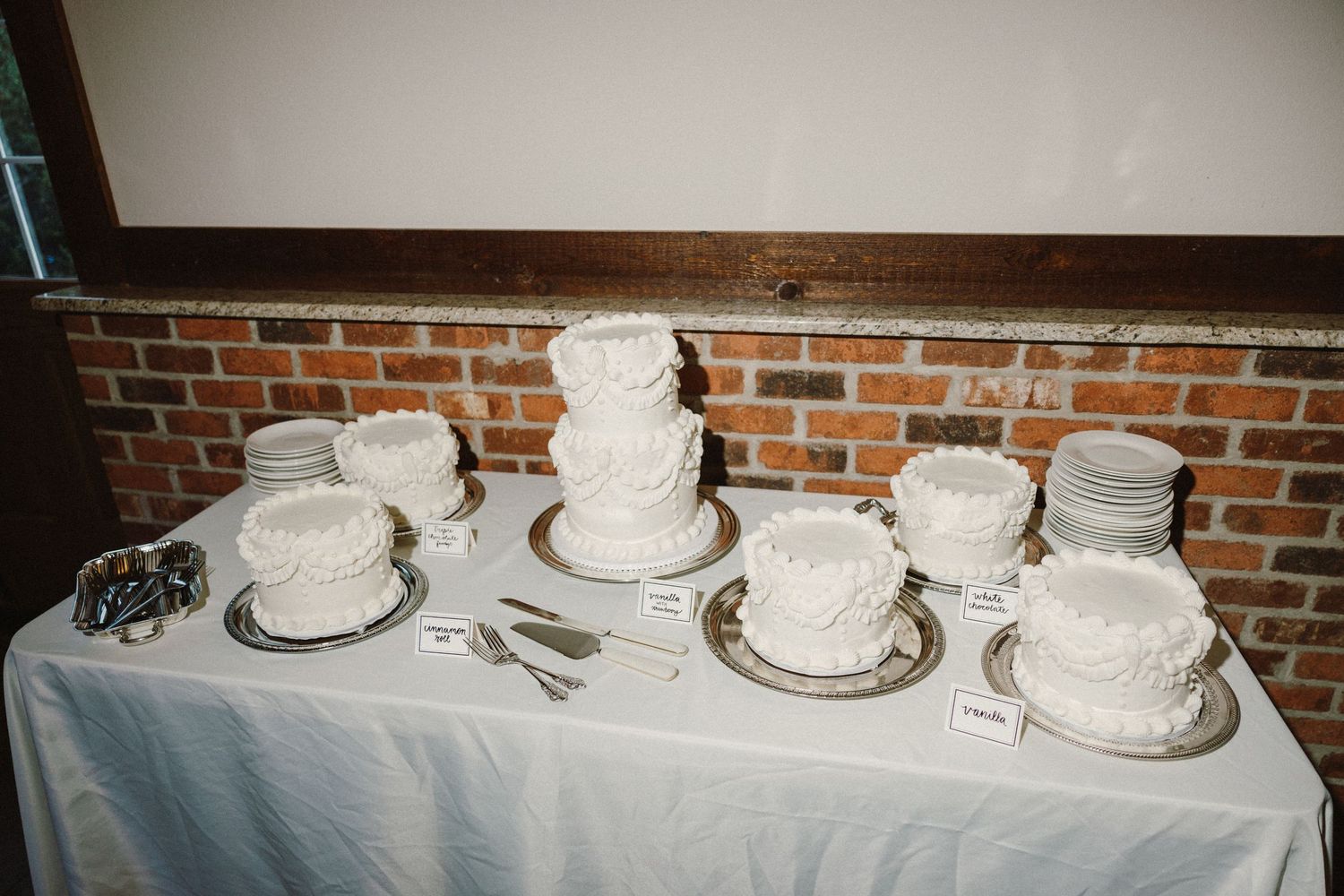 Multiple white wedding cakes displayed on a table with white tablecloth against a brick wall.