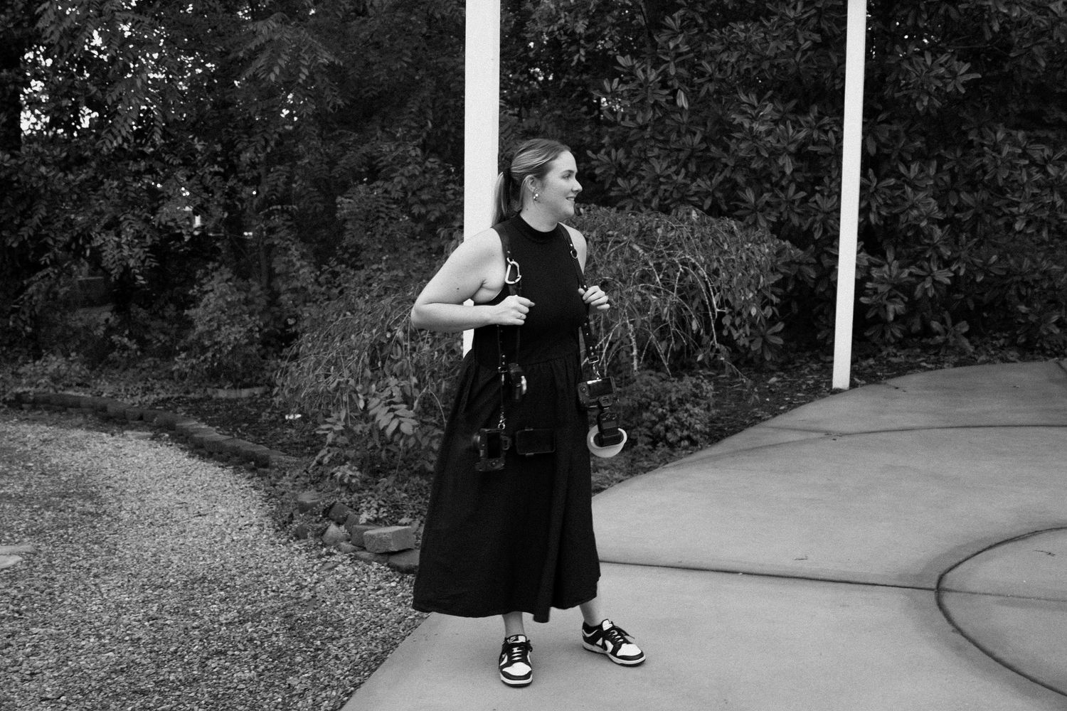 A person in a long black dress and sunglasses stands on a curved path near foliage in black and white photography.