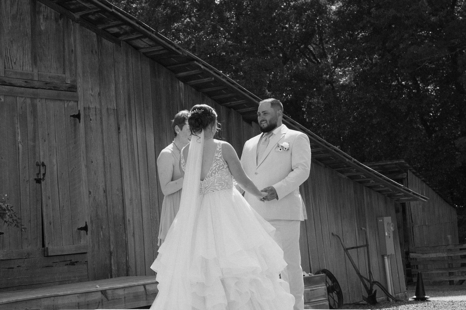 Black and white photo of a couple in wedding attire standing together outside a rustic wooden barn.