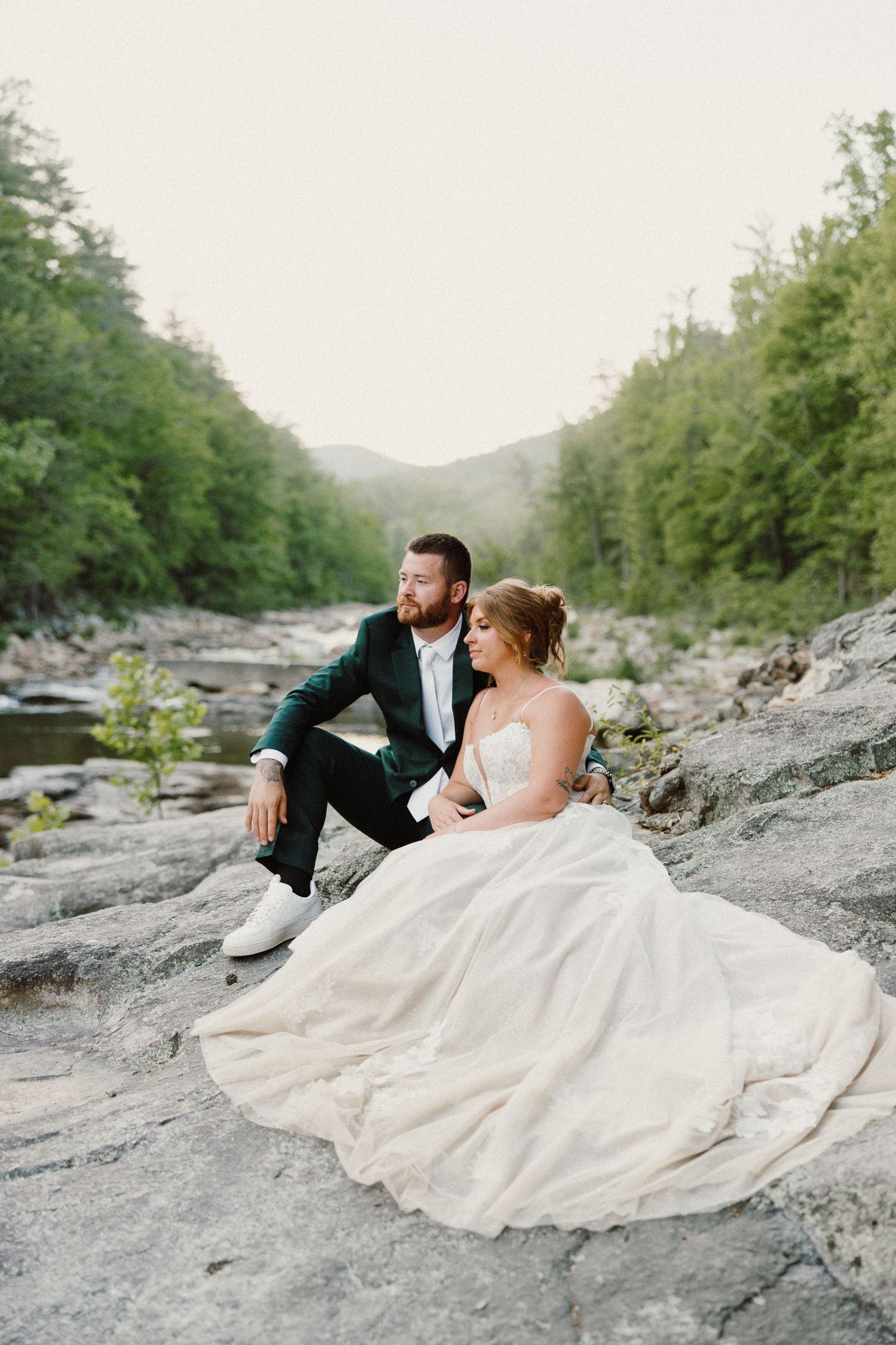 Bride and groom sitting on rocky riverbank surrounded by lush green forest mountains in wedding attire.