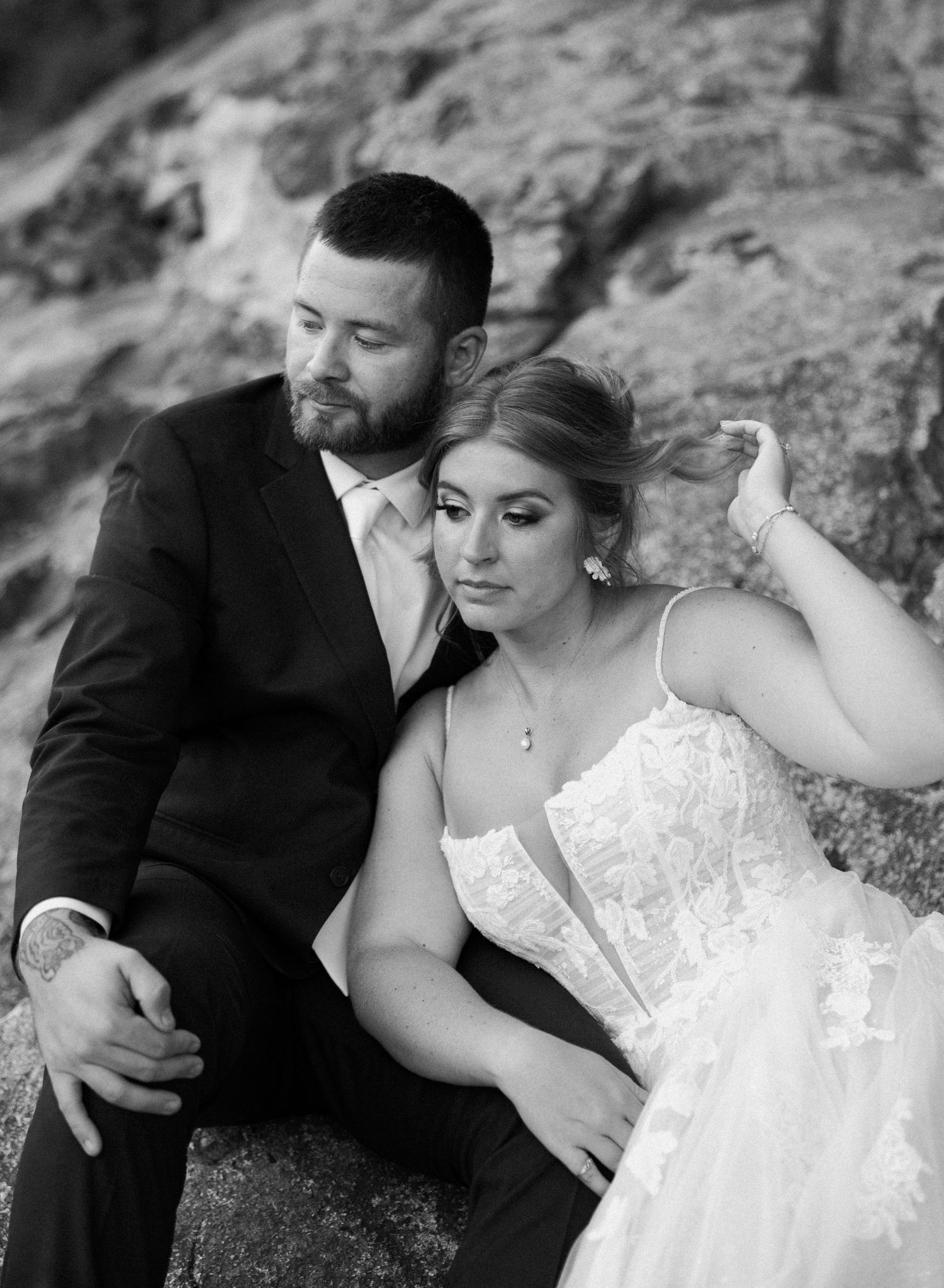 Black and white wedding photo of bride and groom sitting on rocks, groom in suit embracing bride in lace gown.