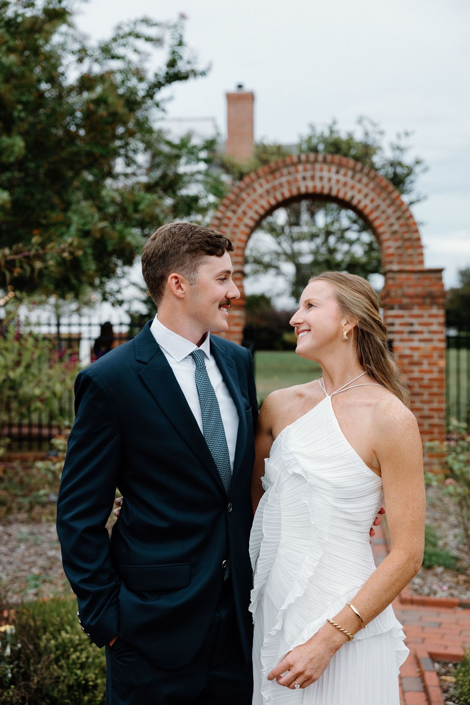 A couple in wedding attire share joyful moments together against a brick wall and garden backdrop.