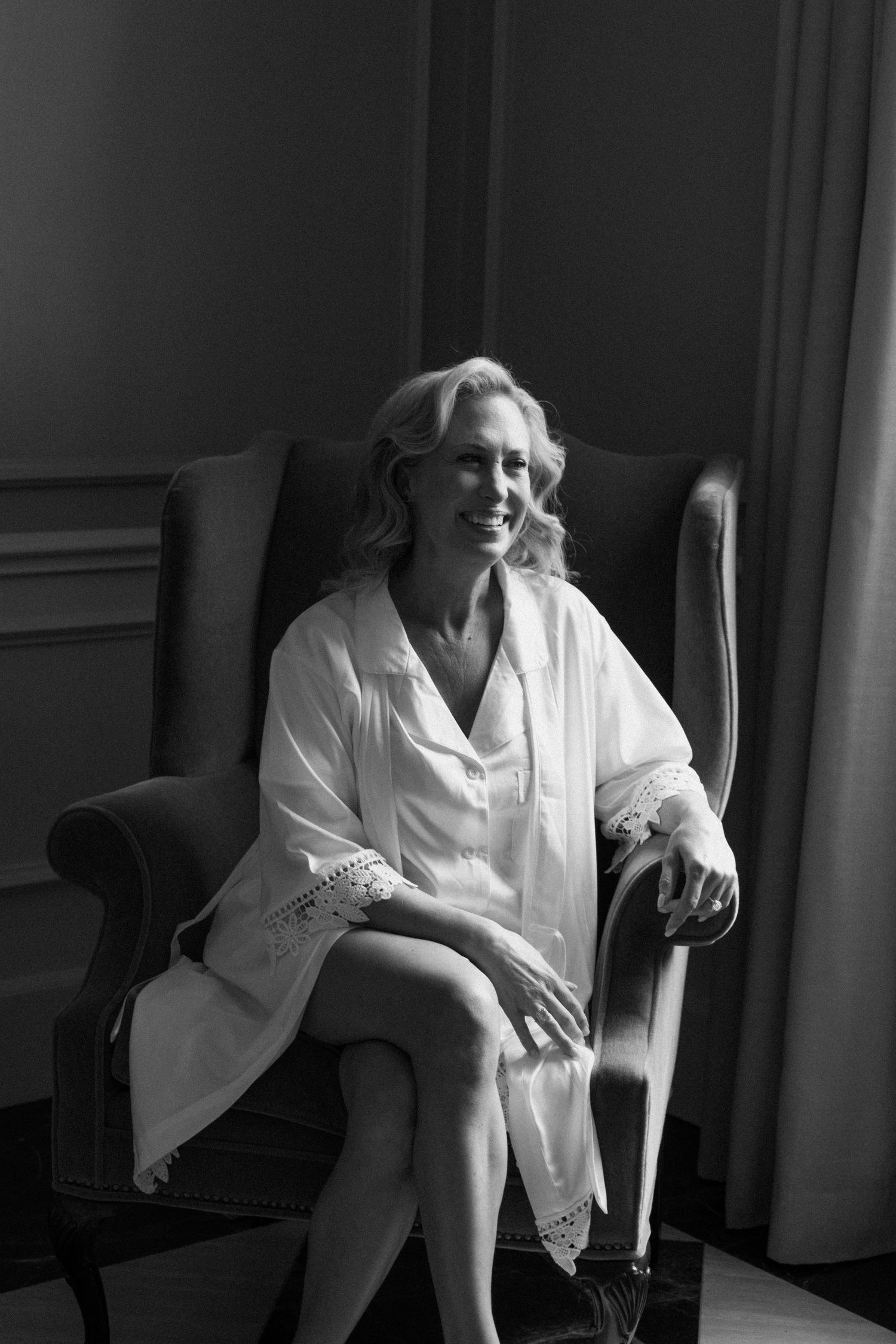 A person in a white robe sits elegantly in an armchair during a black and white photoshoot in vintage style.