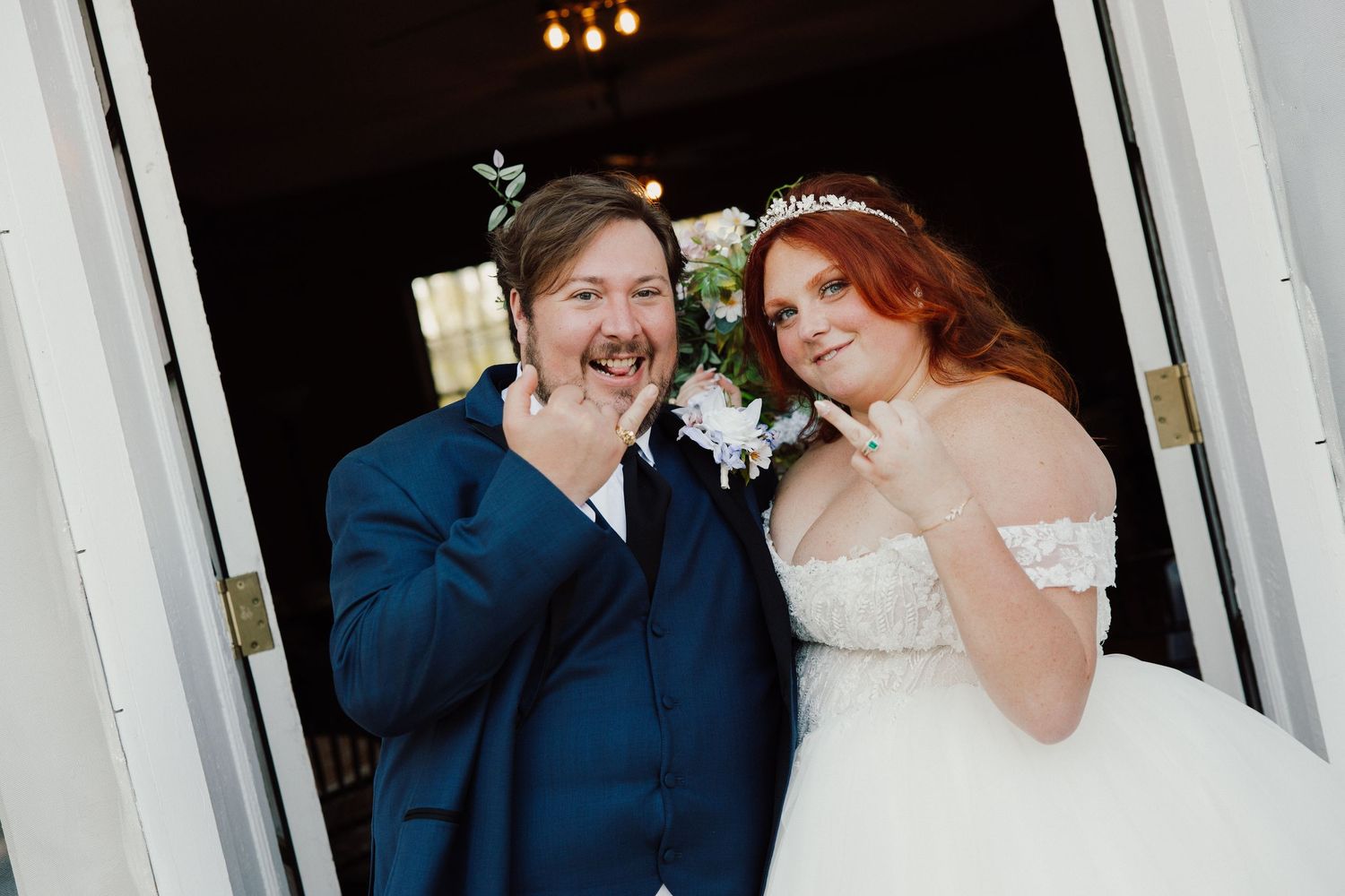 Smiling bride in white off-shoulder gown and groom in navy suit pose together at wedding venue doorway.