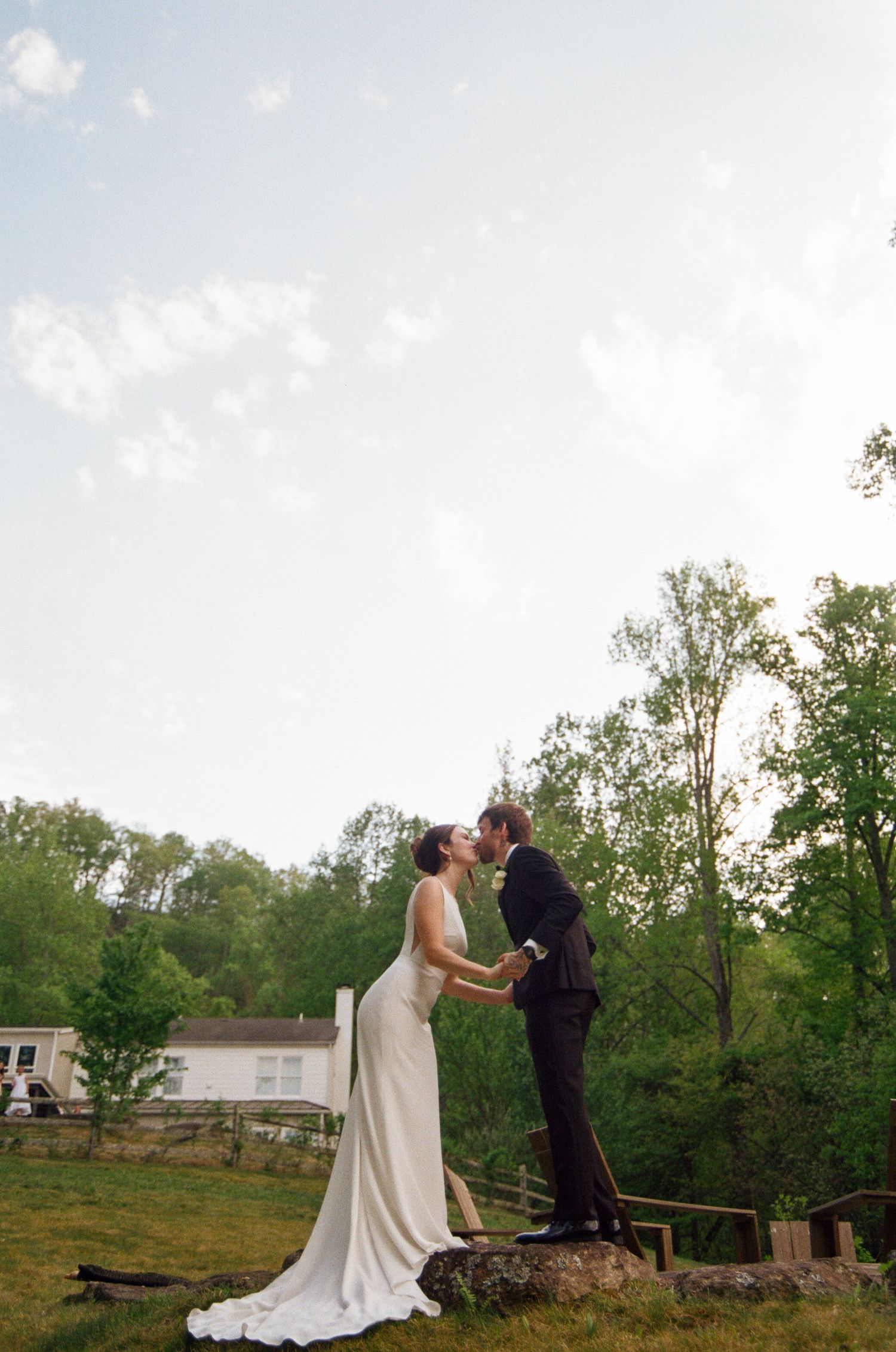 A bride and groom share a kiss while standing on a rock in a lush green outdoor setting on their wedding day.