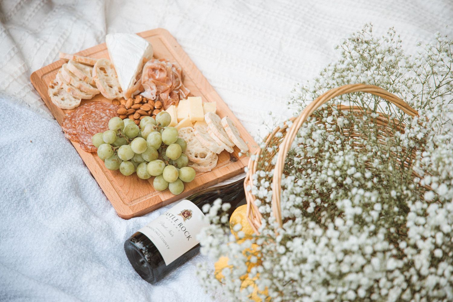 Rustic picnic setup with cheese board, grapes, and baby's breath flowers on white linen.