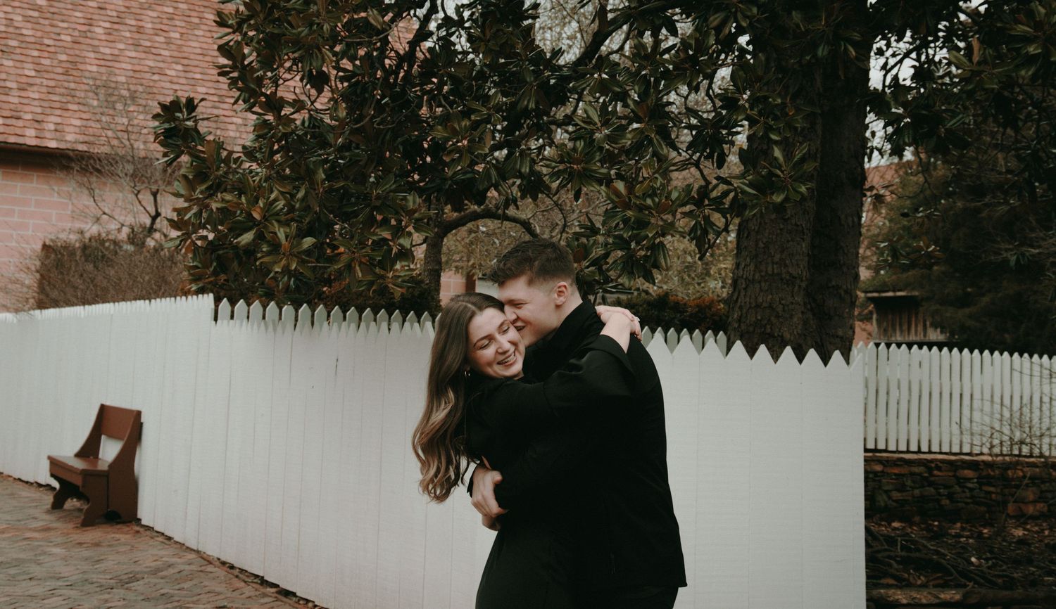 Intimate moment between couple by white picket fence in moody outdoor setting.