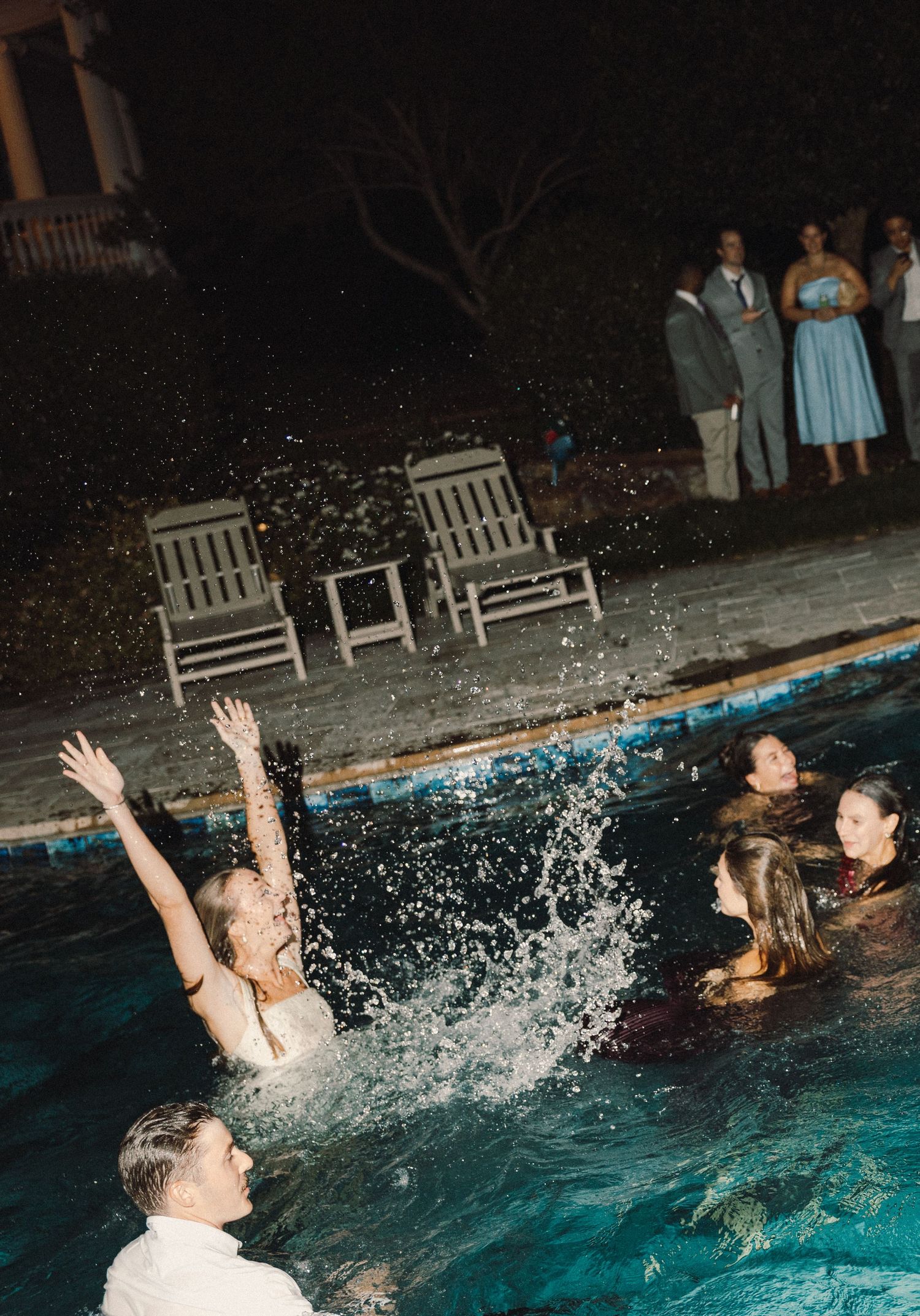 People splashing and playing in an illuminated swimming pool at night with empty chairs visible by the poolside.