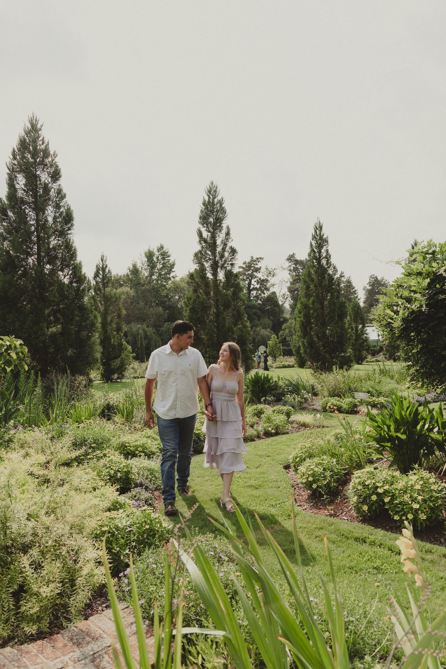 A couple walks hand in hand through a lush garden pathway on a cloudy day.