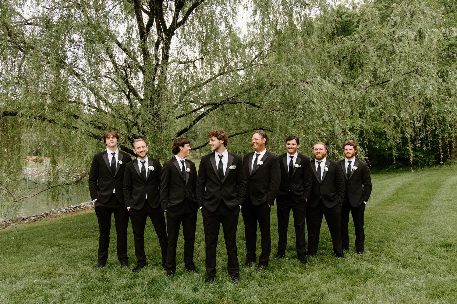 Groomsmen in matching black suits and ties stand on green lawn beneath drooping willow trees at outdoor wedding venue.