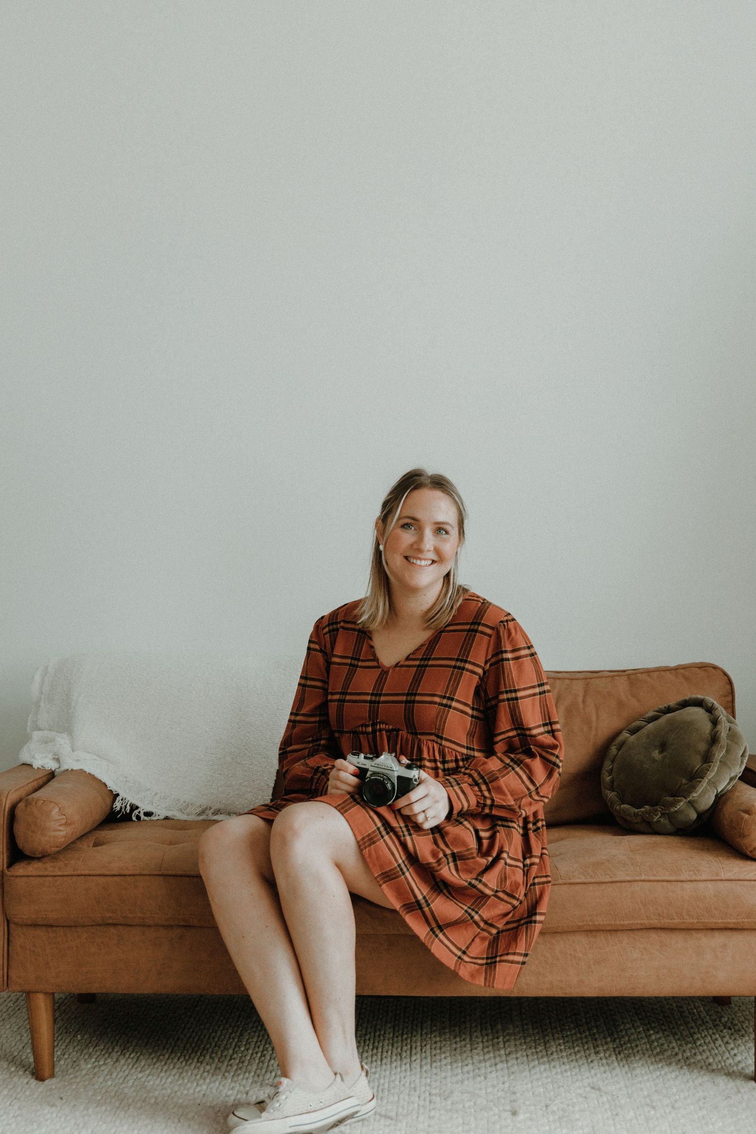 Photographer in plaid dress sitting on vintage brown couch against white wall.