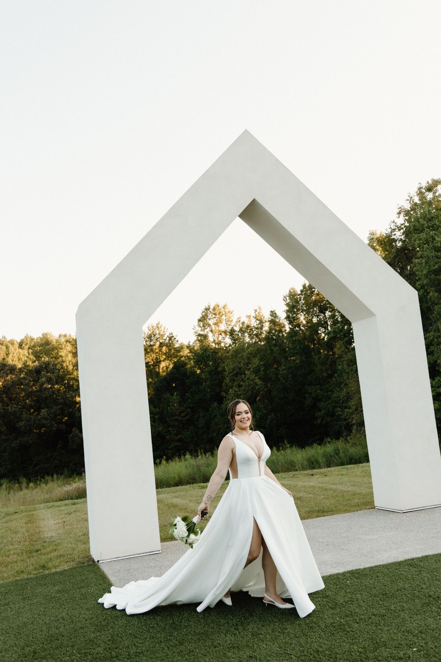 A bride in a flowing white wedding gown poses gracefully against modern white architectural arches on a grassy lawn.