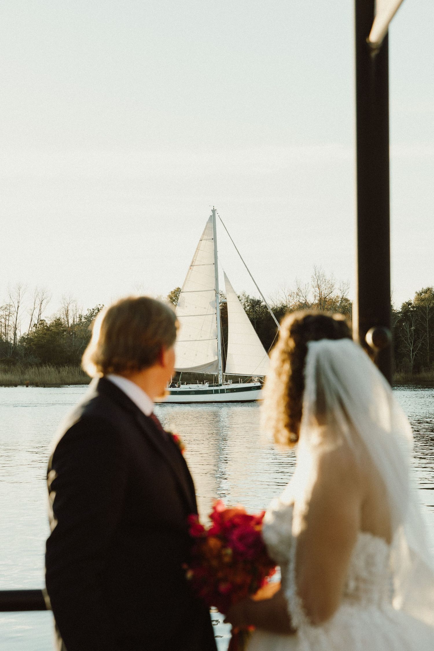 A wedding couple stands by the water at sunset with sailboats in the background.