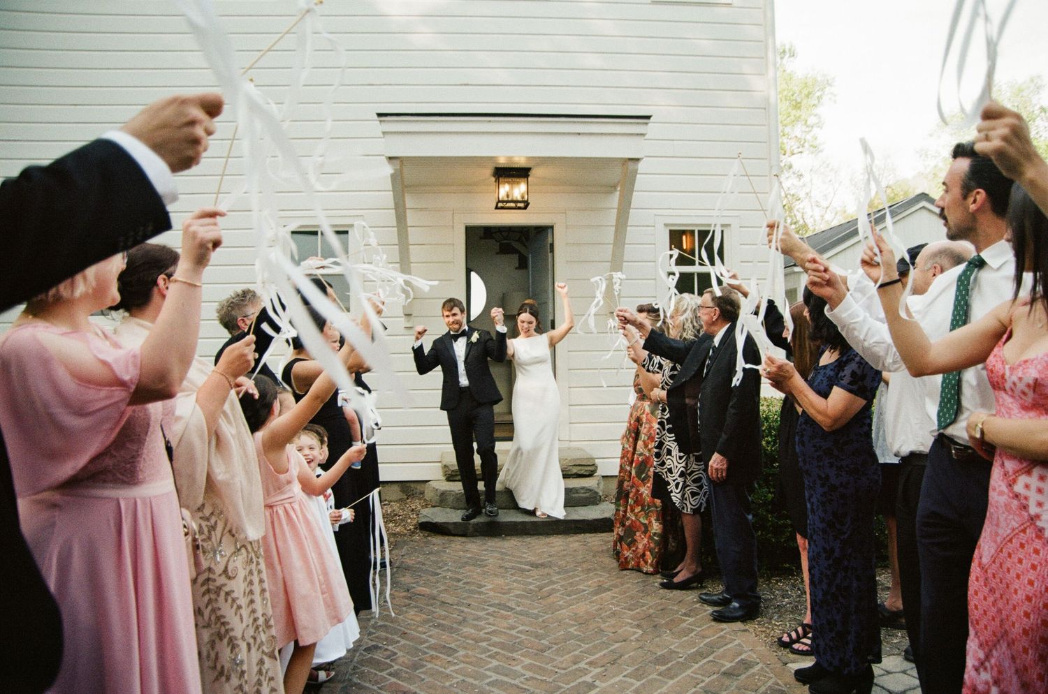 Bride and groom exit wedding chapel as guests wave ribbon wands in joyful outdoor celebration.