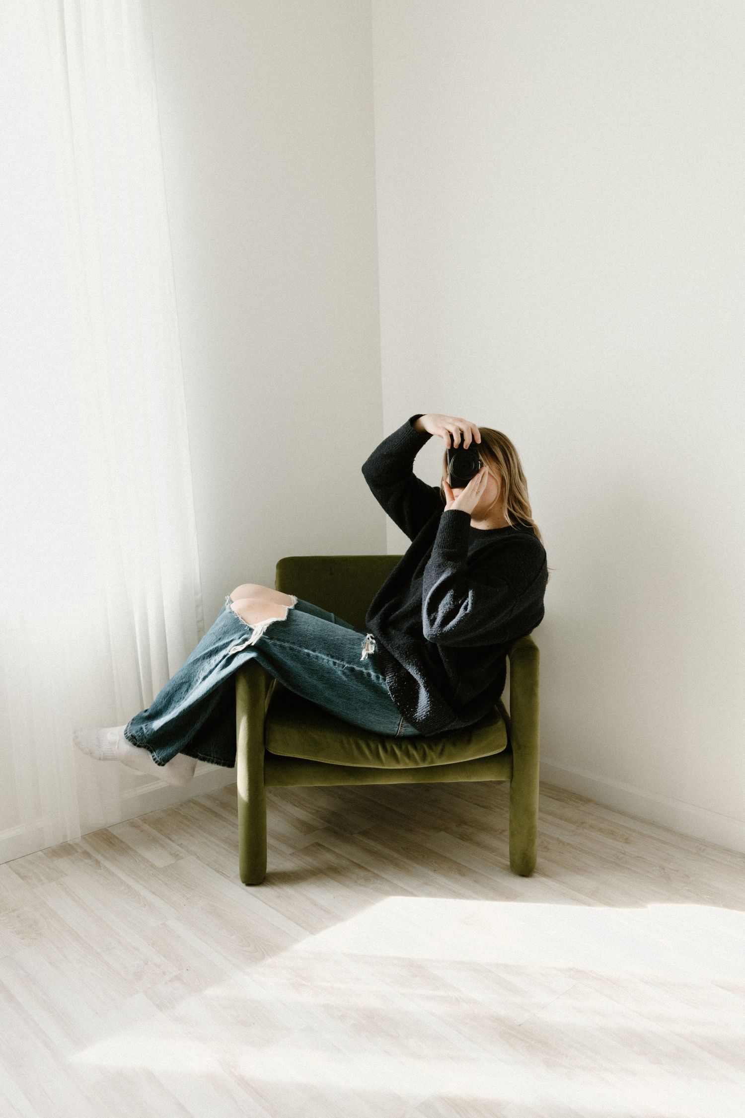 Someone wearing black lounges casually in an olive green armchair against a minimalist white wall.