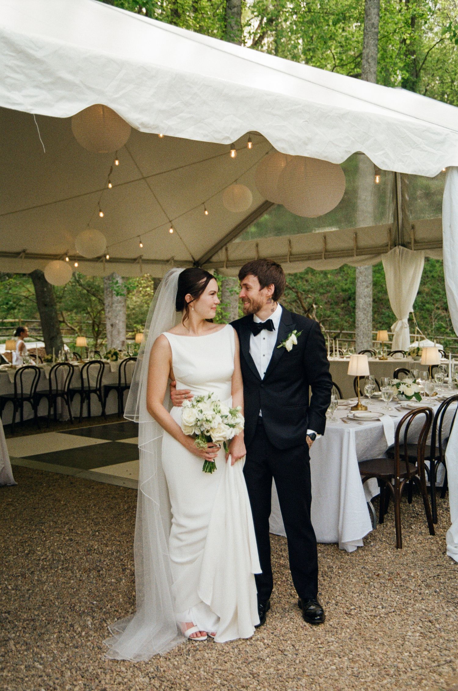 Bride and groom stand together under elegant white wedding tent with string lights and reception tables in wooded outdoor setting.