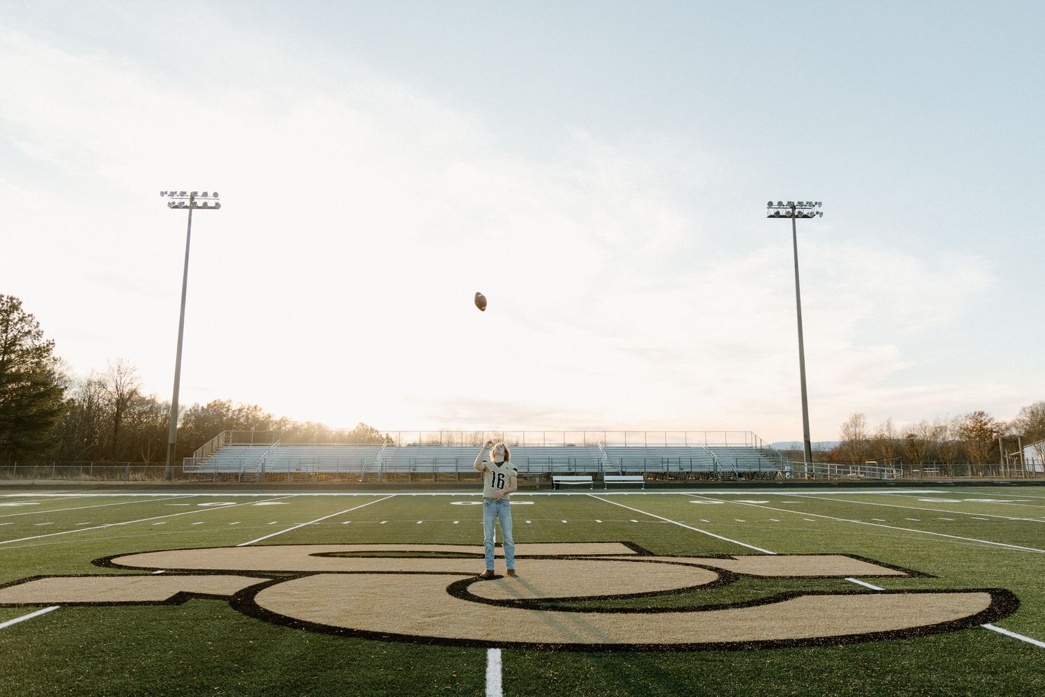 Someone stands on a football field at sunset tossing a ball in the air beneath stadium lights.
