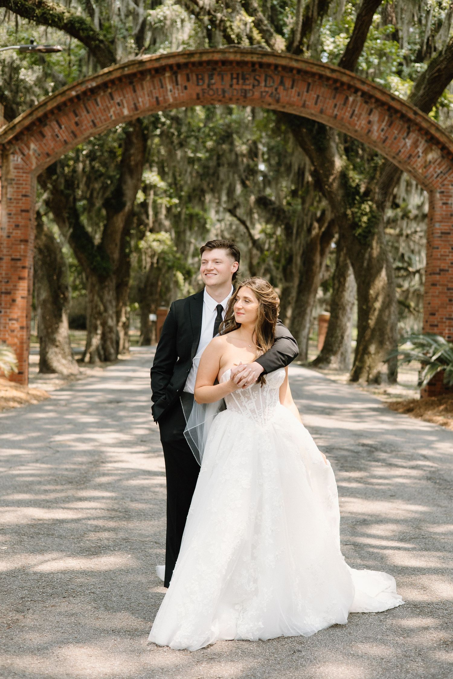 Two newlyweds laugh and dance together on a tree-lined path with Spanish moss hanging from above in romantic sunlight.