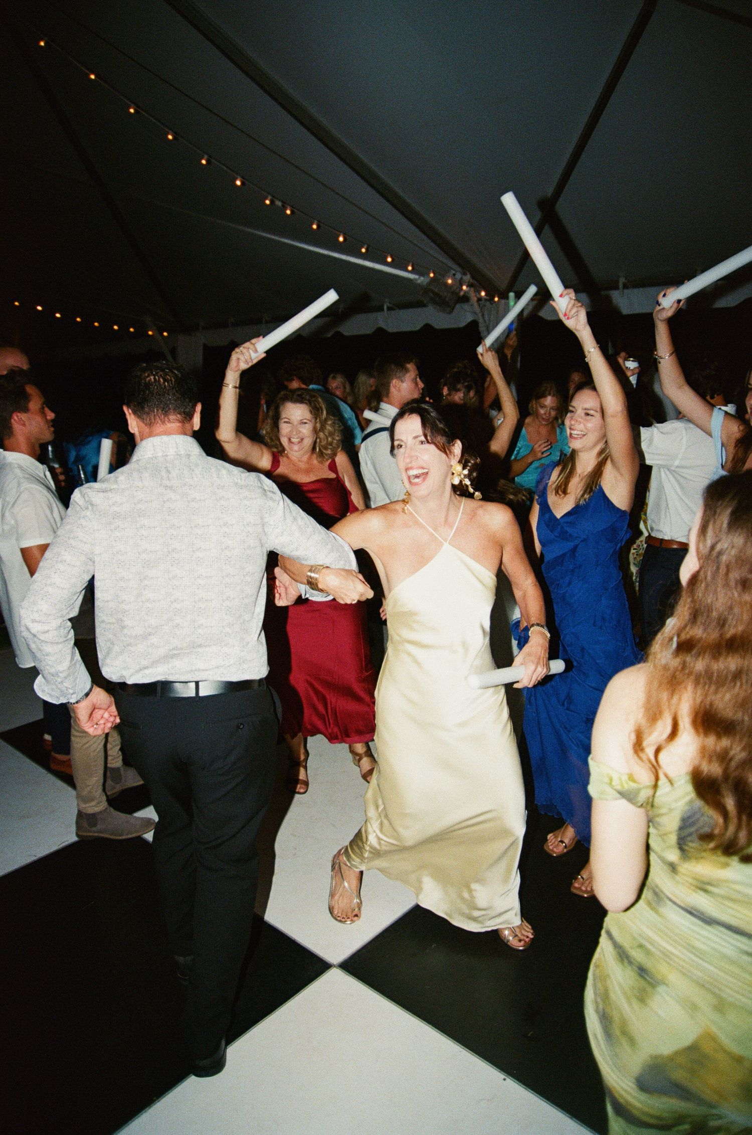 Wedding guests dance and celebrate with light-up foam sticks under string lights at an evening reception party.