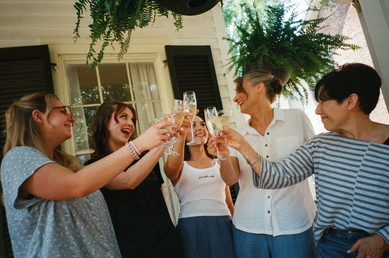 Five women toasting with champagne glasses outdoors, celebrating together under lush green hanging plants.