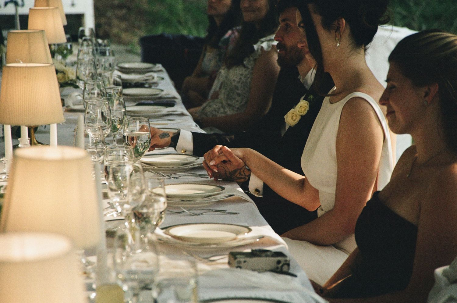 Elegant wedding reception dinner with guests seated at a long table adorned with glassware, candles, and white place settings.