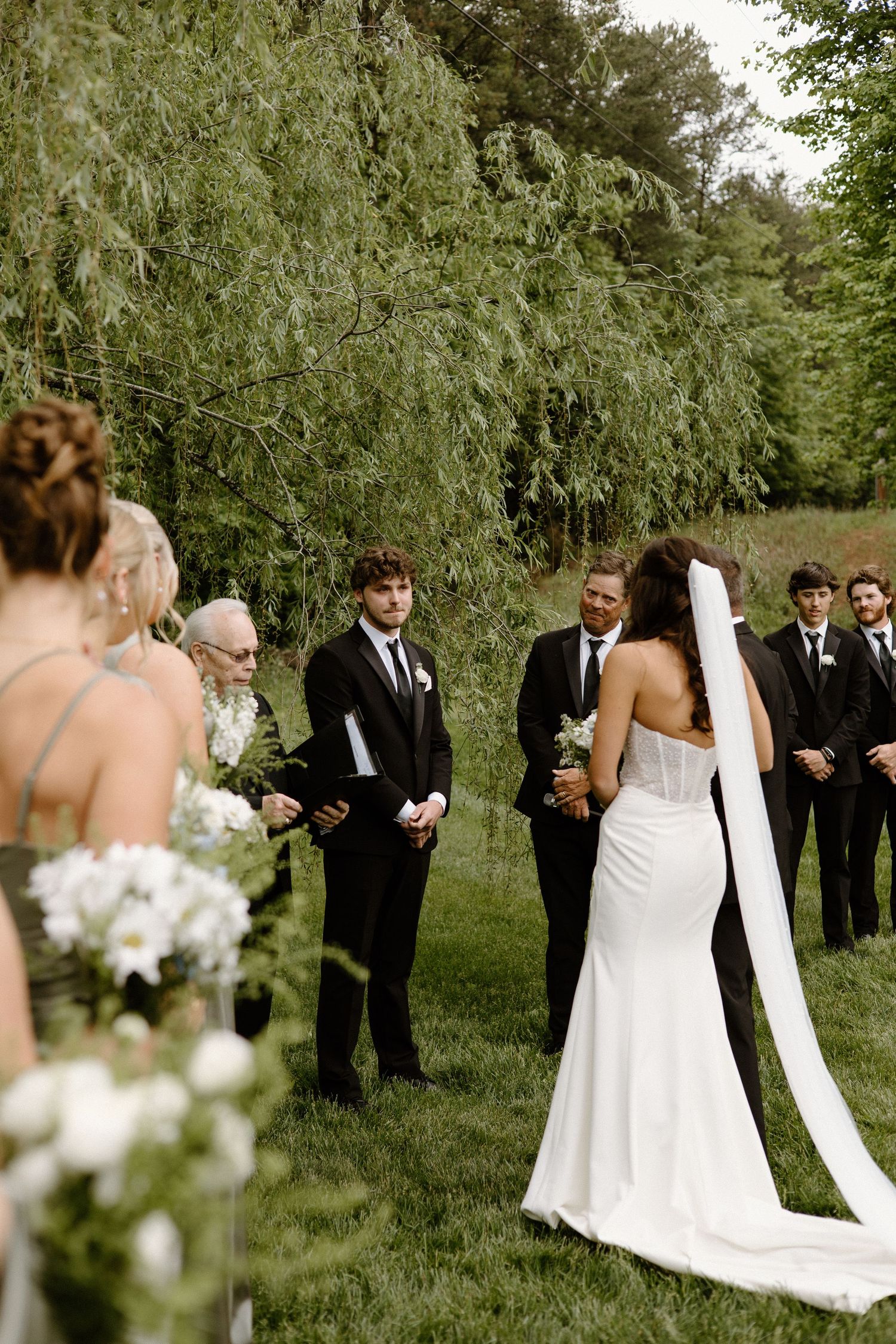 A wedding ceremony takes place outdoors under willow trees with white floral arrangements and attendants in dark suits.
