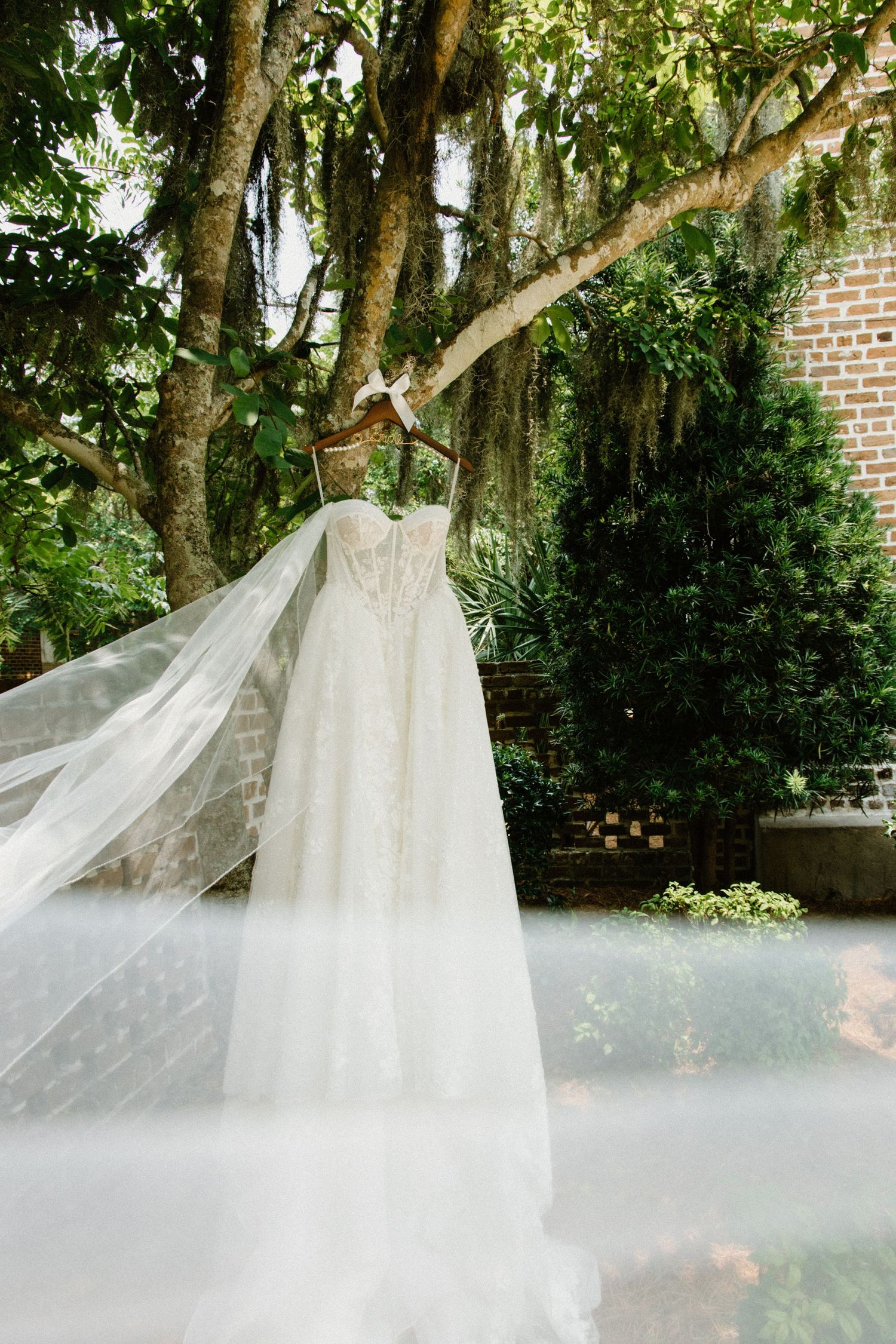 A flowing white wedding veil blows gracefully in the wind against a backdrop of trees and brick walls.