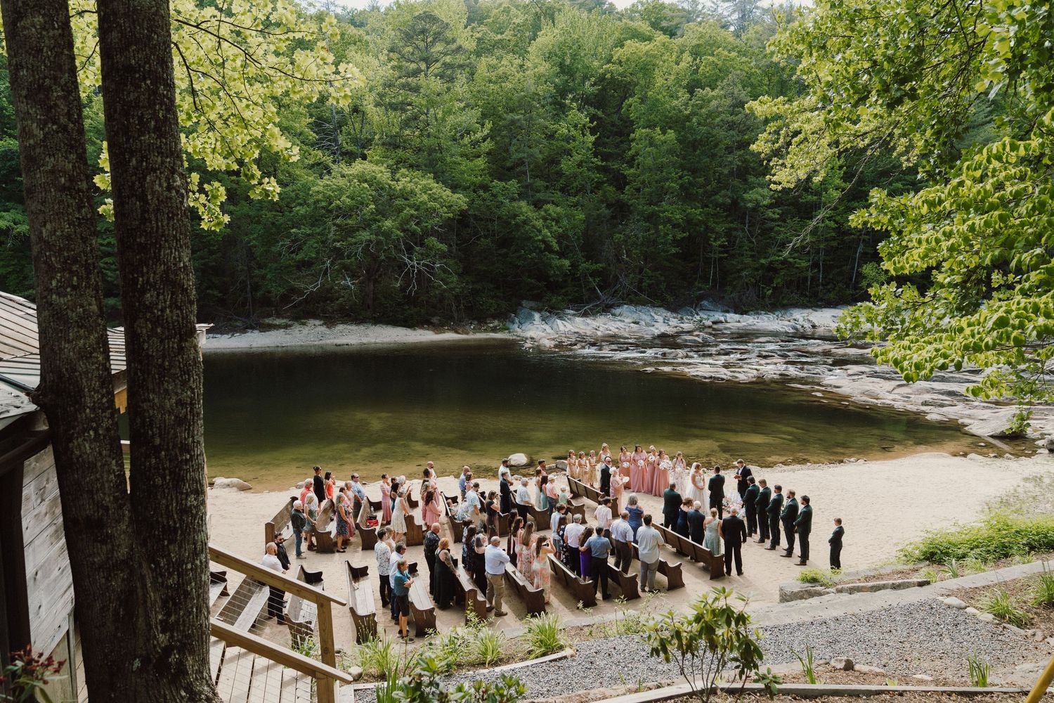 Outdoor wedding ceremony on a sandy riverbank surrounded by lush green forest with guests seated in rows.