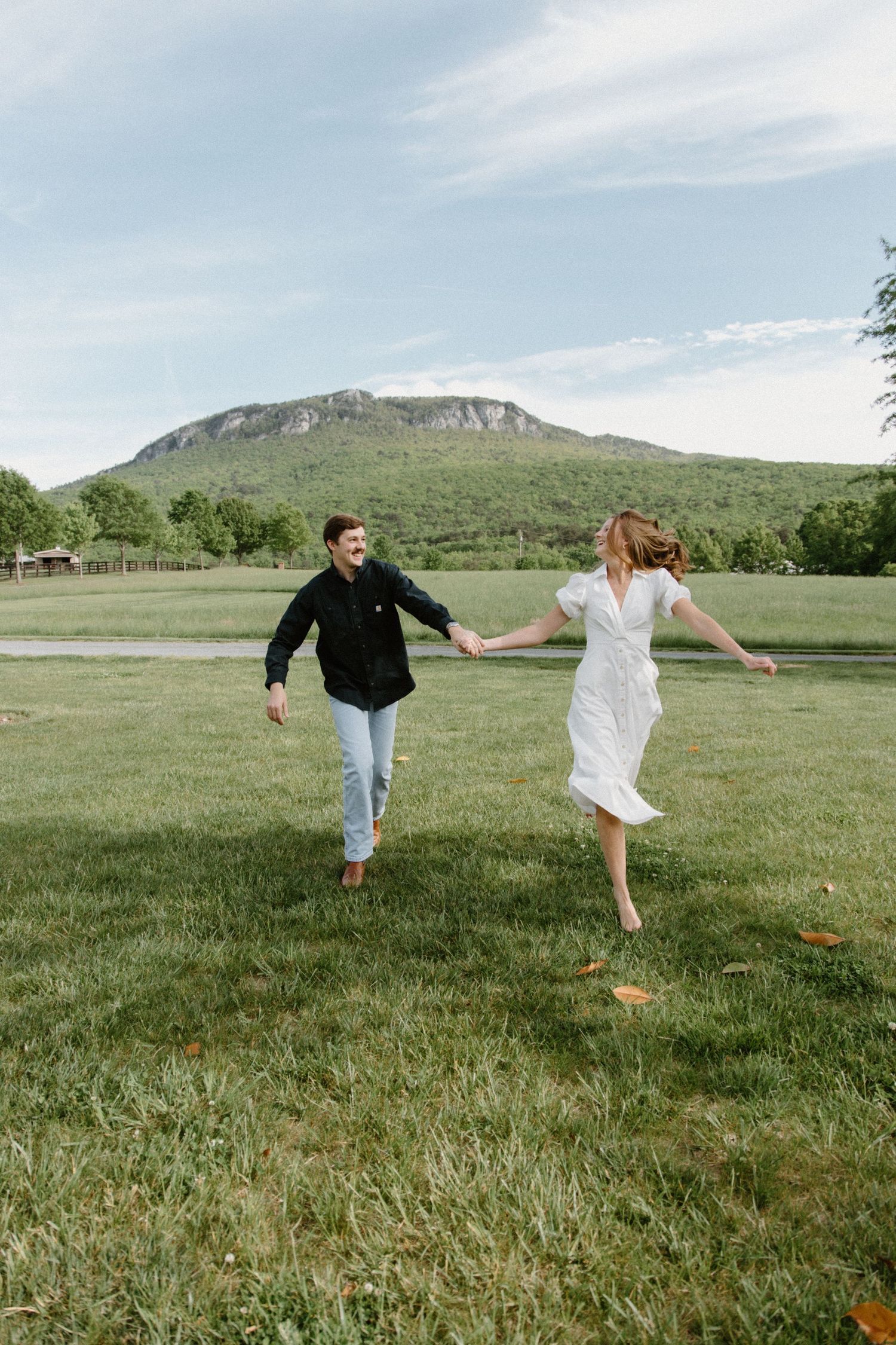 Two people dance and spin playfully in a green meadow with mountains and blue sky in the background.