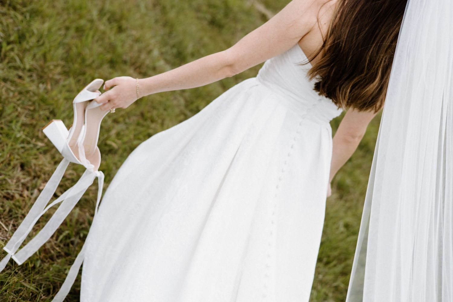 A wedding dress and veil flow in the wind as a bride holds her white high heels outdoors on grass.