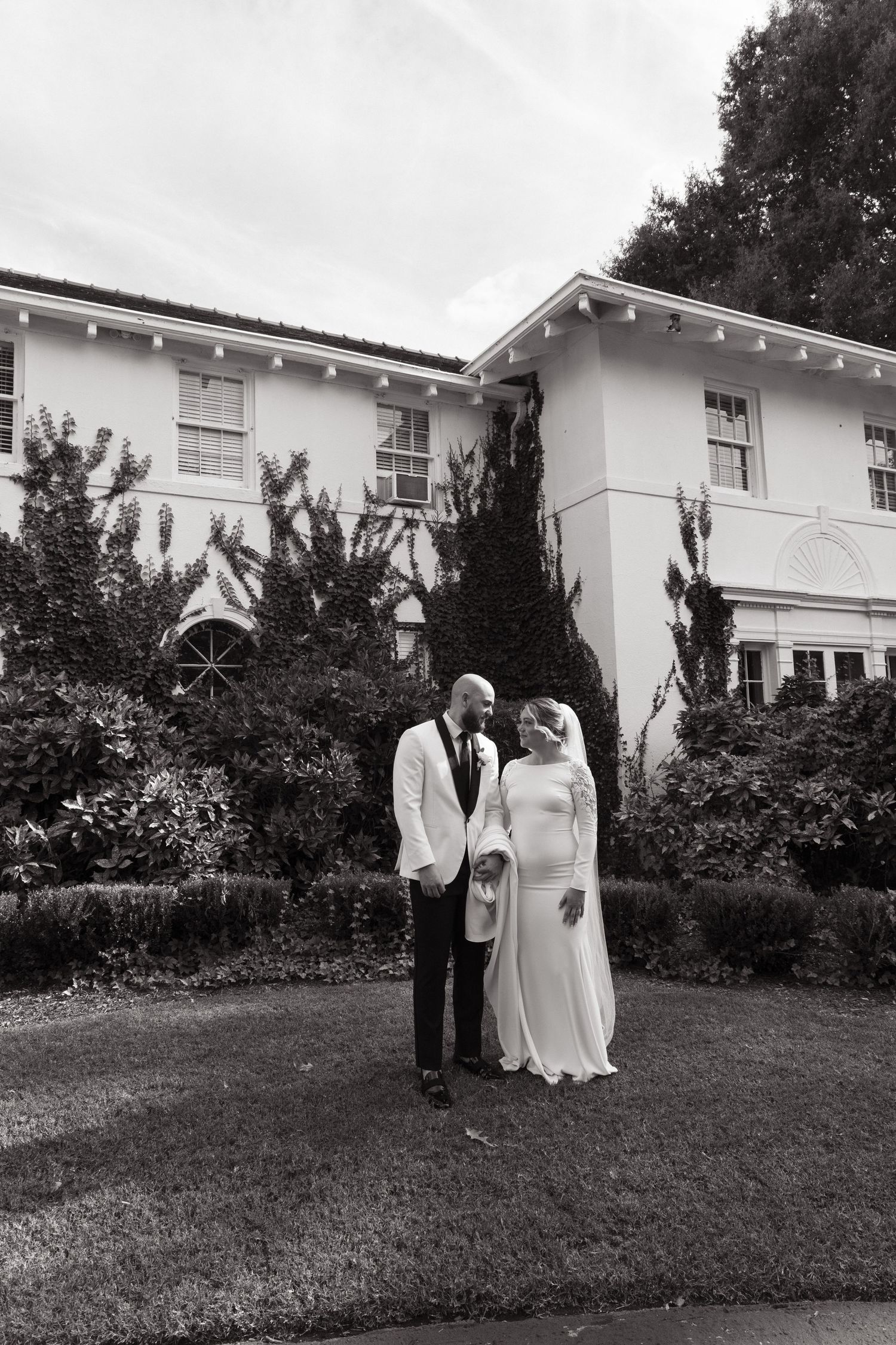 A couple stands together on a manicured lawn in front of a grand white two-story house.