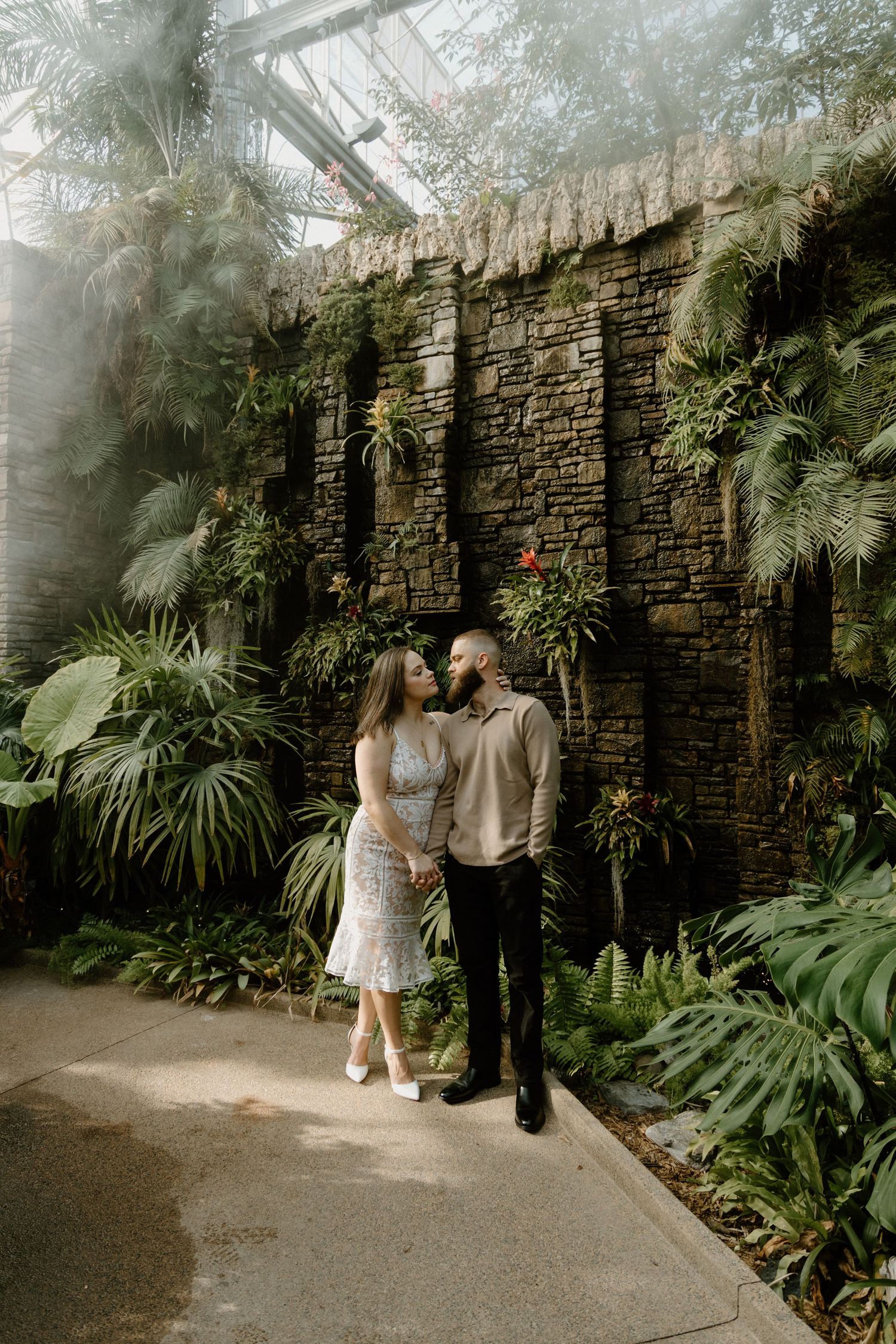 A couple poses together in a lush greenhouse setting with stone walls and tropical plants as their backdrop.