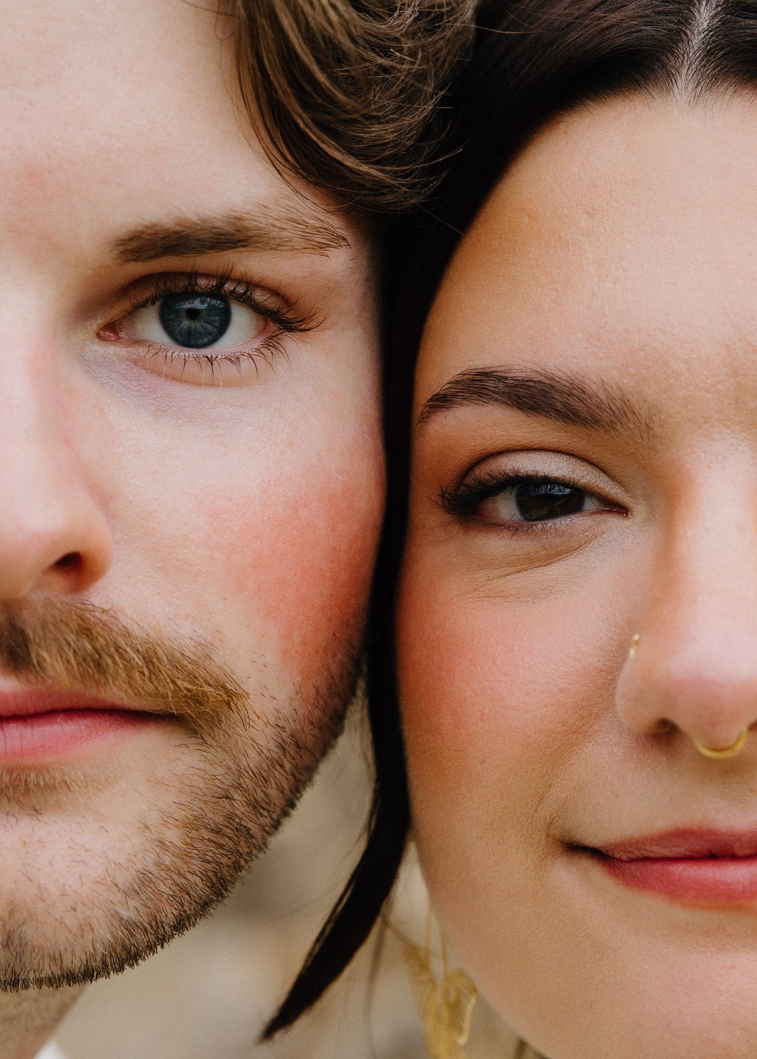 Close-up portrait of a young couple with dark hair, blue eyes, and a nose ring, cheek to cheek.